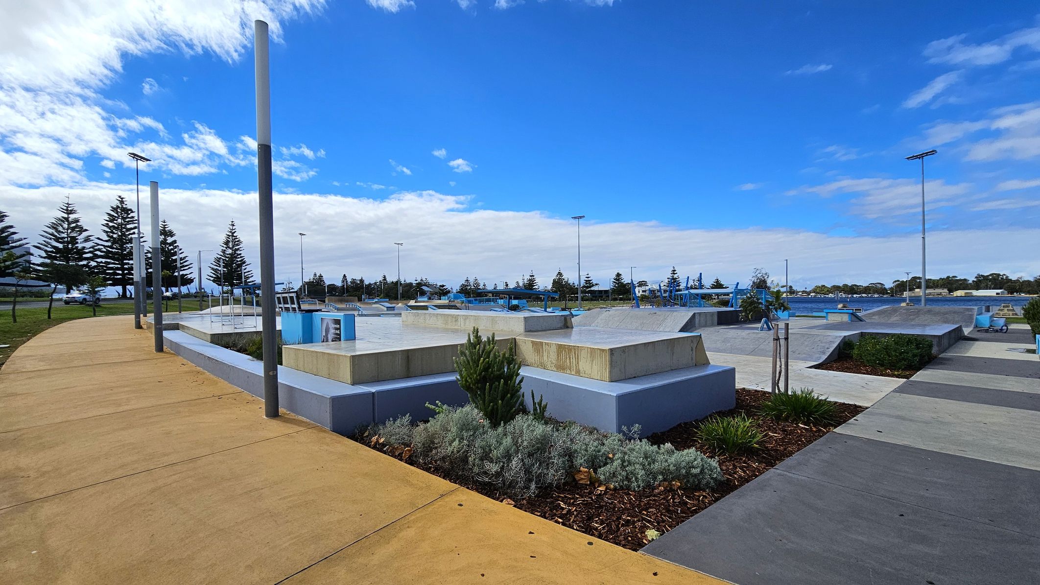 A skate park with various ramps and platforms is in the centre. On the left, a curved, tan-coloured path runs alongside tall metal poles and a row of trees. On the right, there are more ramps and a grey pathway. In the foreground, there are landscaped areas with shrubs and small plants. In the background, more trees and a clear blue sky are visible.