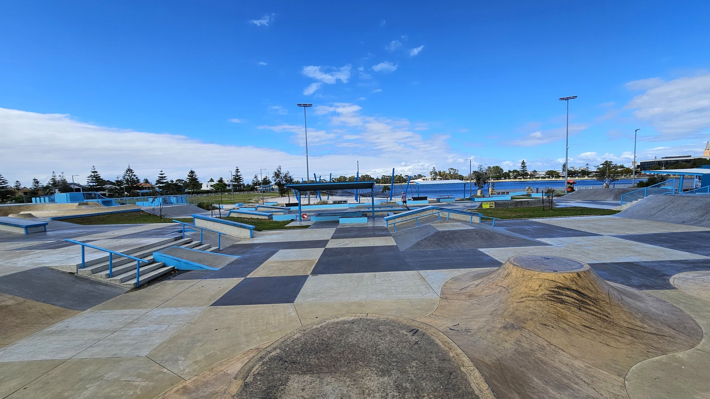 A skate park with various ramps, rails, and platforms in shades of blue and grey. The ground is a patchwork of different coloured concrete sections. In the background, there are trees and a body of water. Several tall light poles are visible, and a few people are scattered throughout the park. The area is open and spacious, with a clear view of the surroundings.
