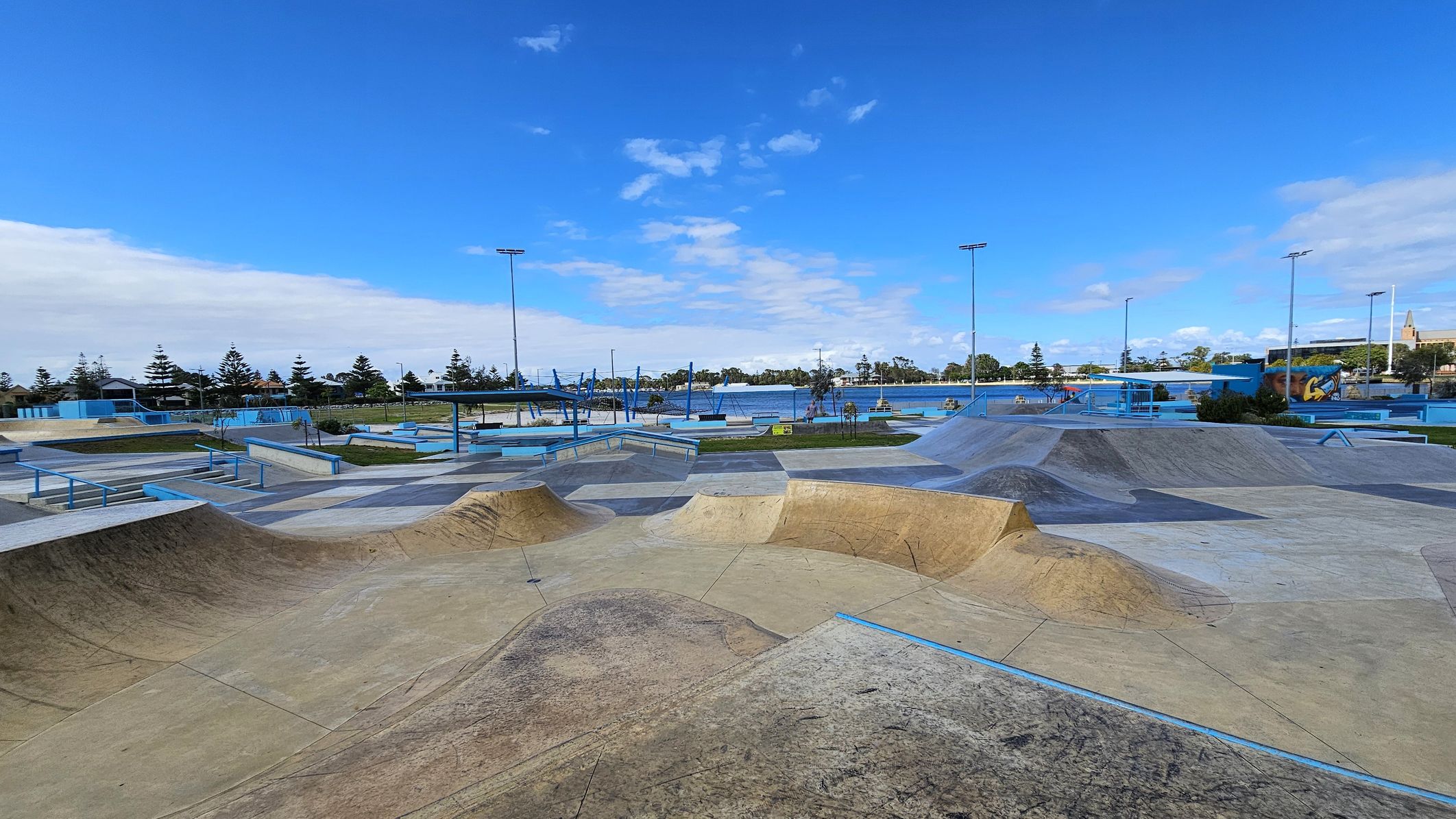 A skate park with various ramps and rails is in the foreground, featuring a mix of concrete and metal structures. The middle ground has more skate features and a few trees. In the background, there are tall light poles, a body of water, and a row of trees. A mural is visible on the right side. The sky is clear with some clouds.