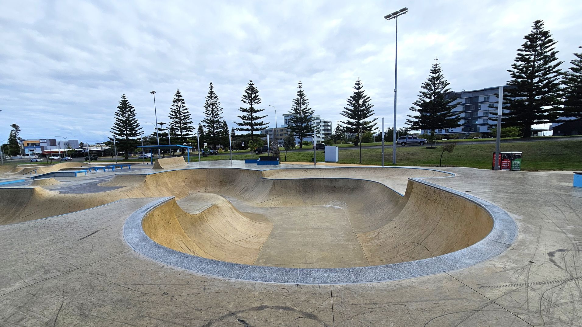 A skate park with smooth concrete surfaces and curved ramps in the foreground. In the background, there are tall pine trees lining the area, with a few modern buildings visible beyond them. The ground is marked with various lines and scuffs from skateboards. There are street lamps and a few benches scattered around the park. A couple of bins are positioned on the right side near the trees.