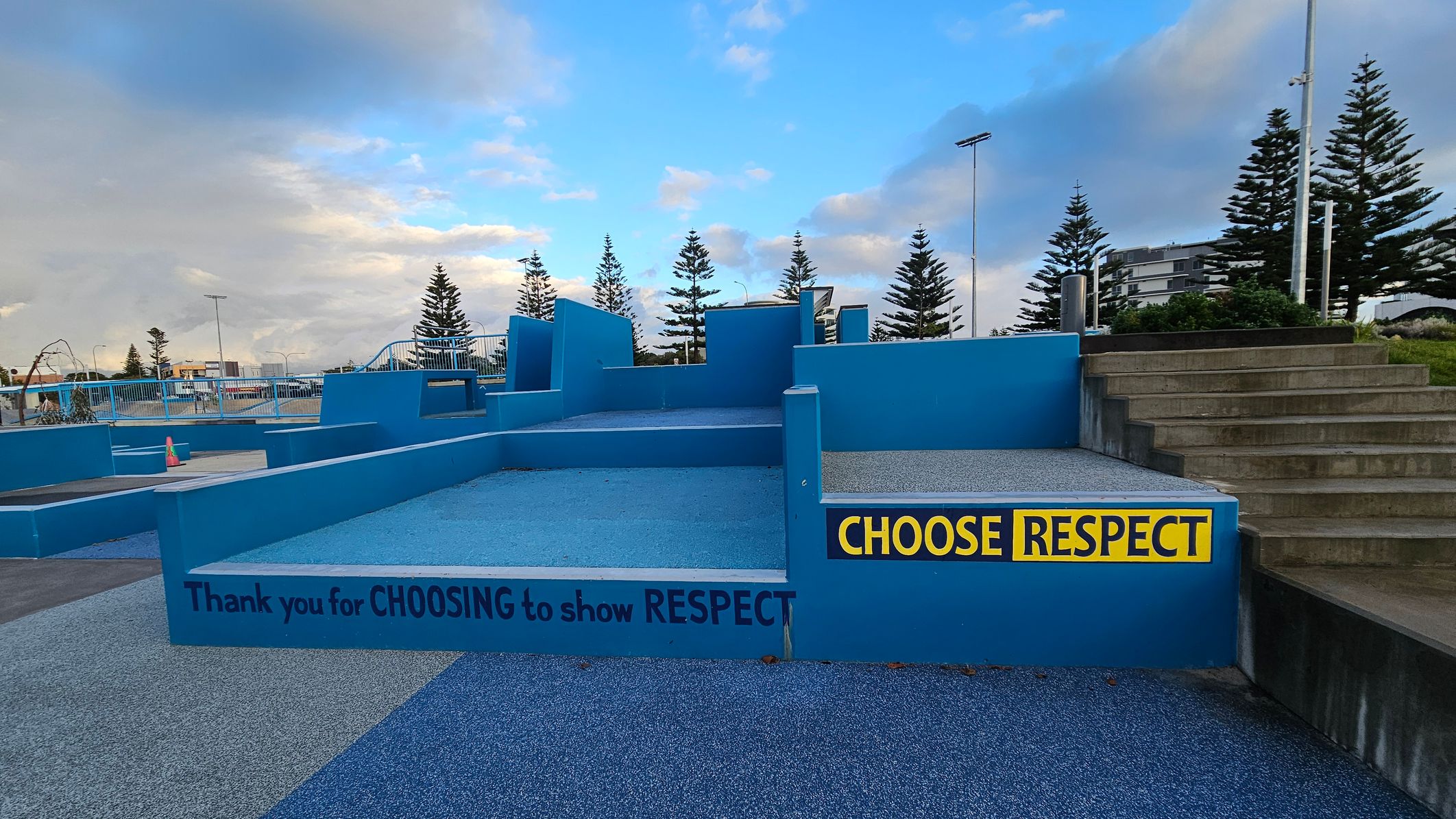 A blue concrete structure with multiple levels and steps is in the centre. On the right, a set of grey steps leads up to a grassy area. A yellow and black sign reads "CHOOSE RESPECT." Another message on the structure says, "Thank you for CHOOSING to show RESPECT." In the background, there are tall pine trees and a few buildings. The ground is covered with a textured blue surface, and there are railings and a traffic cone visible on the left.