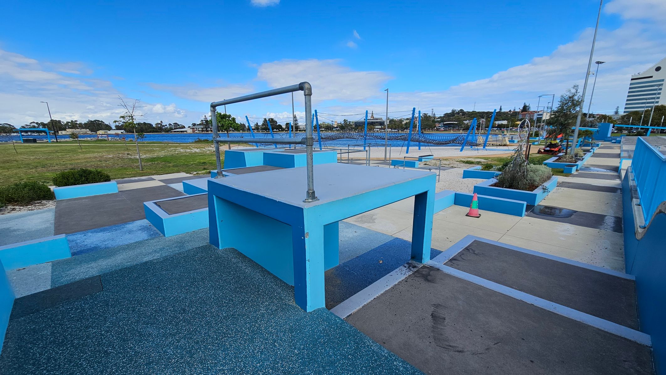 A parkour training area with various blue structures and platforms. In the foreground, a large blue platform with a metal bar on top is visible. To the right, there are more platforms and a pathway with a traffic cone. In the background, a grassy area and a body of water can be seen, along with trees and a few buildings. The ground is a mix of textured surfaces and concrete, and the area is open and spacious.