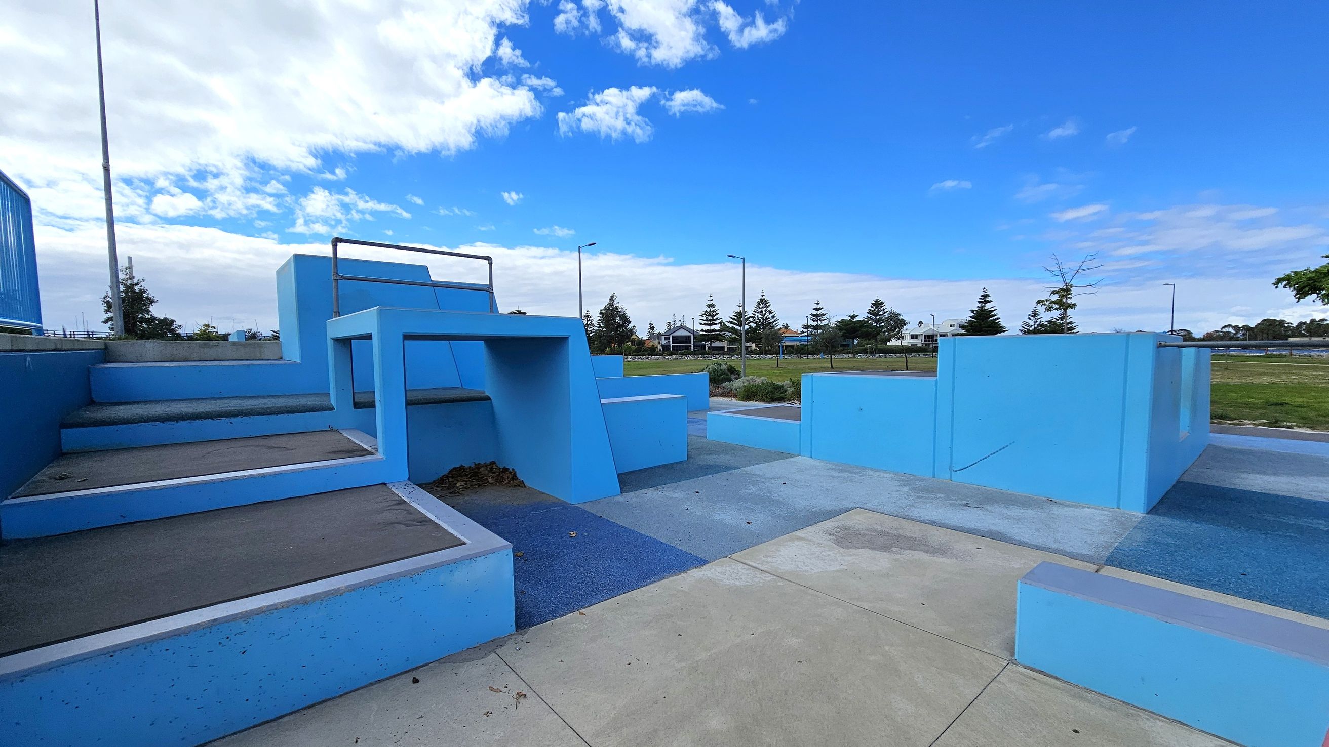 A parkour training area with various blue structures, including steps and platforms, is in the foreground. The ground is a mix of concrete and textured surfaces. In the background, there are trees, a grassy area, and a few buildings. Street lamps line the path, and the sky is partly cloudy.