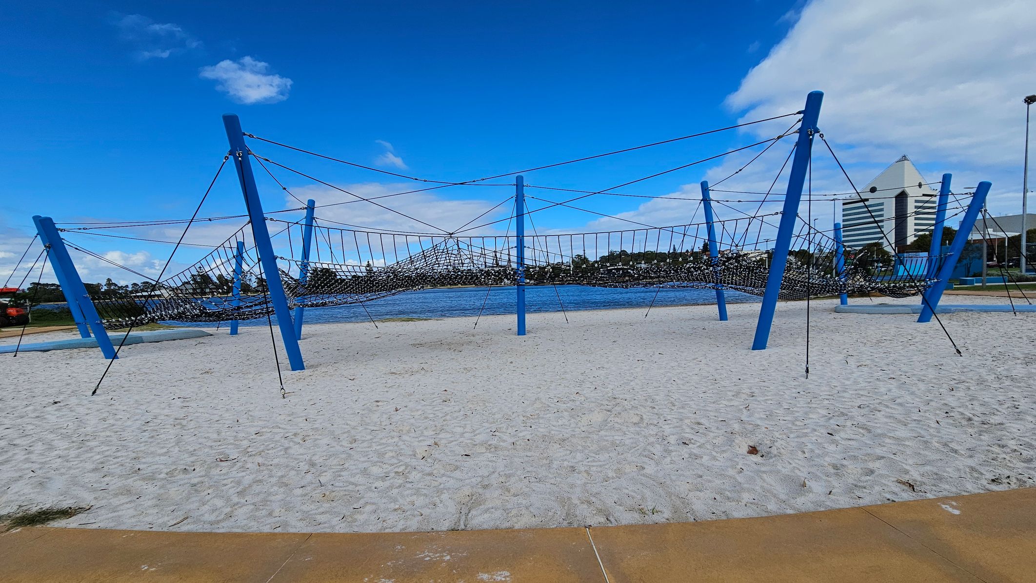 A playground structure with blue poles and a netted climbing area is set on a sandy surface. In the background, there is a body of water with trees lining the opposite shore. To the right, a tall, triangular-roofed building is visible. The foreground shows a paved area bordering the sand.