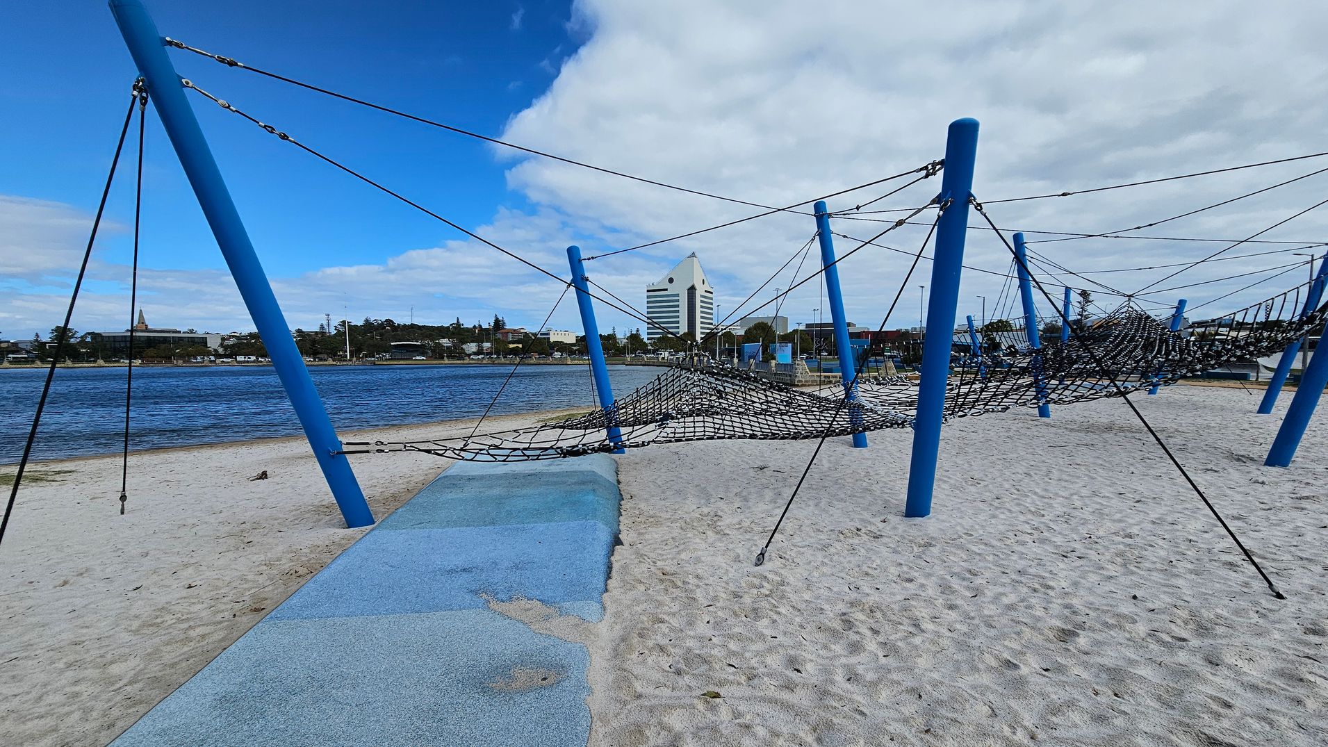 A sandy playground with blue poles supporting a net climbing structure. The foreground has a blue rubber matting area. In the background, a body of water is visible with a cityscape beyond, including a distinctive white triangular-roofed building. The sky is partly cloudy.