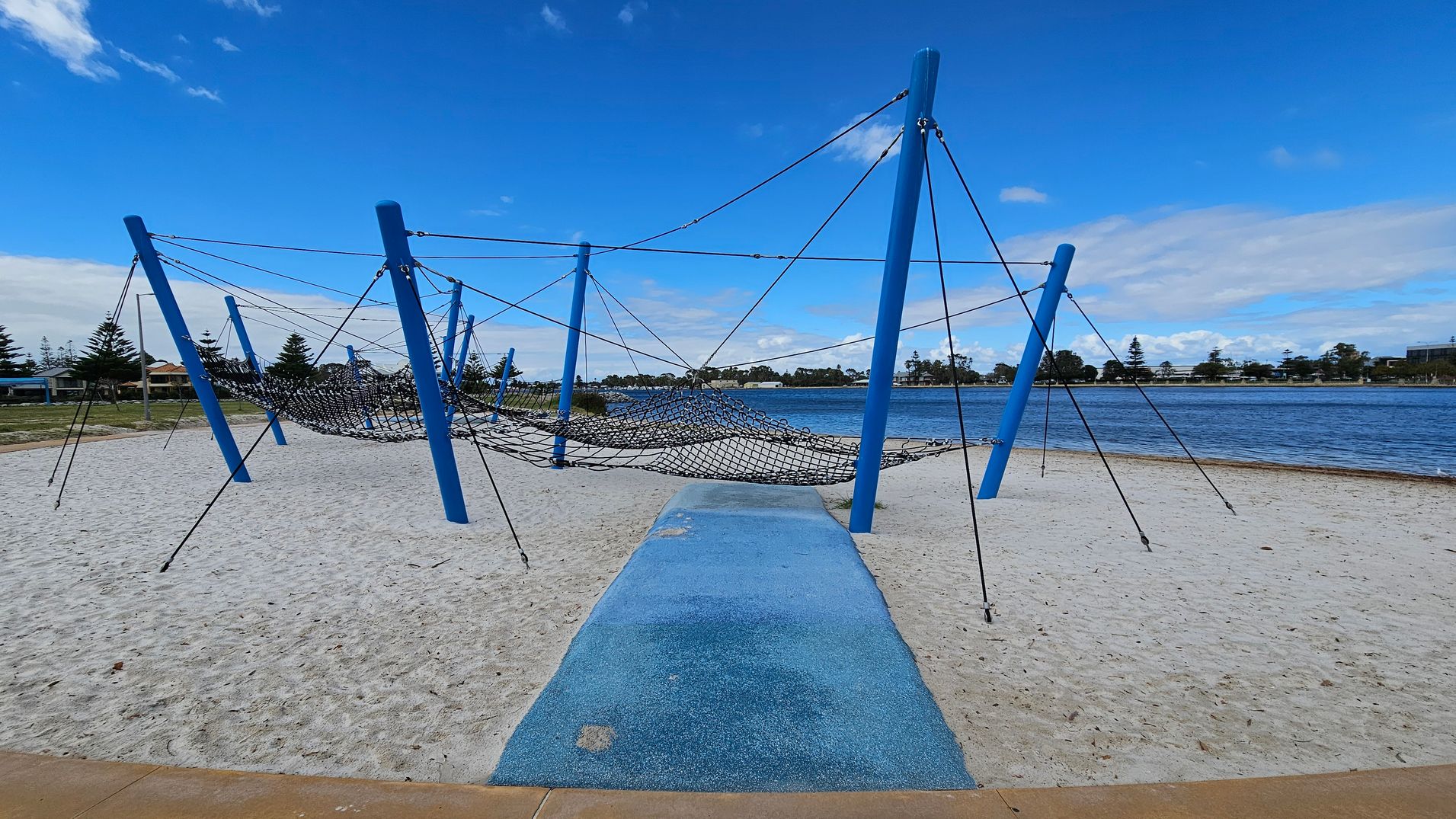 A playground structure with blue poles and a netted climbing area is set on a sandy surface. A blue pathway leads to the structure. In the background, there is a body of water with a shoreline lined with trees and buildings. The sky is clear with a few clouds.