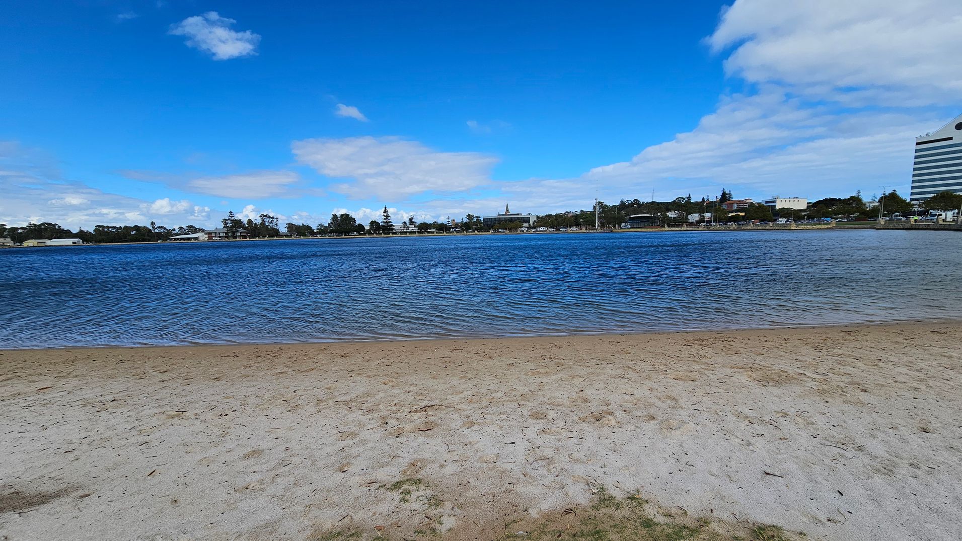 A sandy beach in the foreground leads to a body of water with gentle ripples. Across the water, a distant shoreline features a mix of trees and buildings, including a tall structure on the right. The sky is mostly clear with a few scattered clouds.