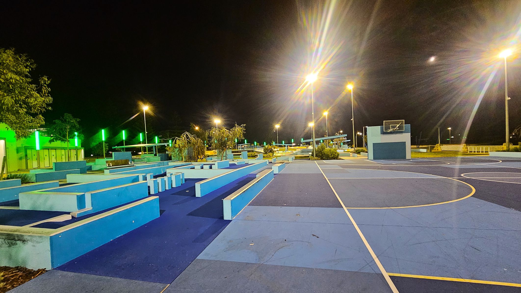 A night-time view of a sports area with a blue and purple basketball court on the right. The court features a hoop with a backboard. On the left, there are geometric blue structures, possibly for seating or parkour. Green lights illuminate the background, with trees and additional structures visible. Tall streetlights cast a warm glow over the scene. The ground is a mix of blue shades, and the sky is dark.