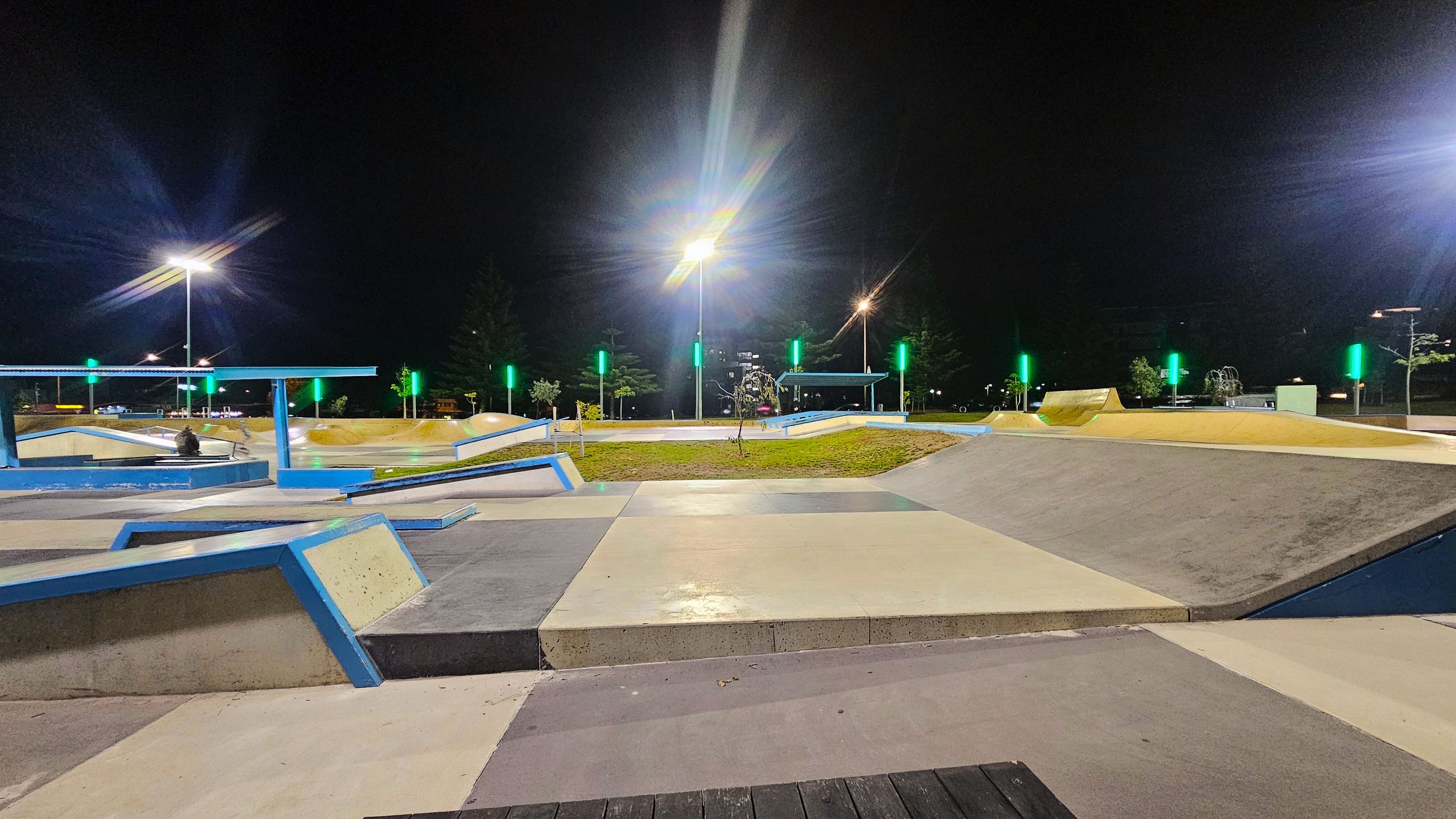 A well-lit skate park at night with various ramps and platforms. The foreground features a smooth concrete surface with blue-edged ramps. In the middle, there are grassy areas and more ramps, with several tall lights illuminating the scene. The background shows trees and additional park structures, with green lights accentuating the area. The sky is dark, indicating nighttime.