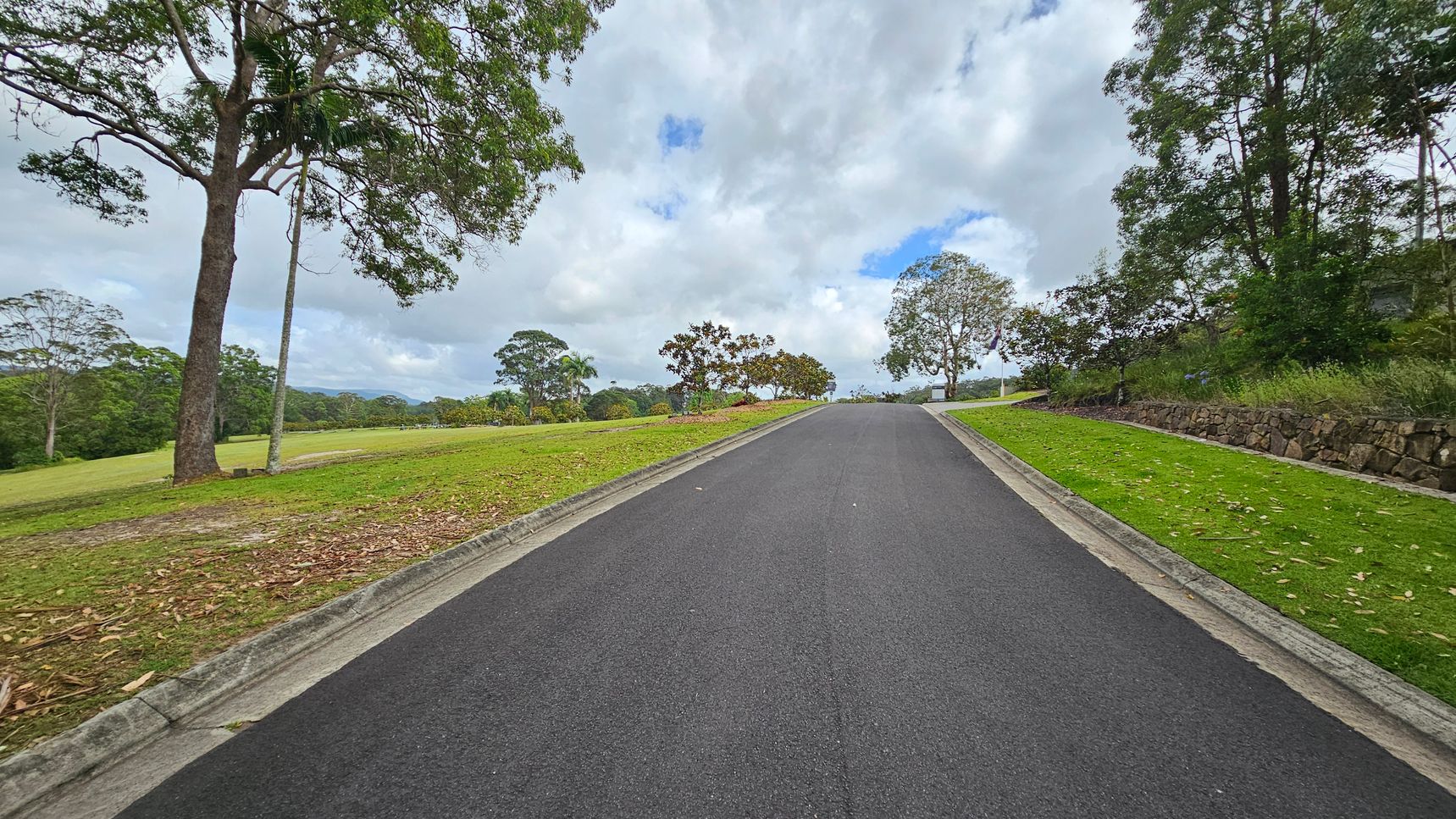 A paved road leads uphill, flanked by tall trees and greenery on both sides. On the left, a sign reads "Kulangoor Cemetery Est. 1907" near a large tree. On the right, a small metal box is attached to a concrete pillar. The road is bordered by grass and shrubs, with speed limit signs visible. The sky is overcast, and the area is surrounded by dense foliage.