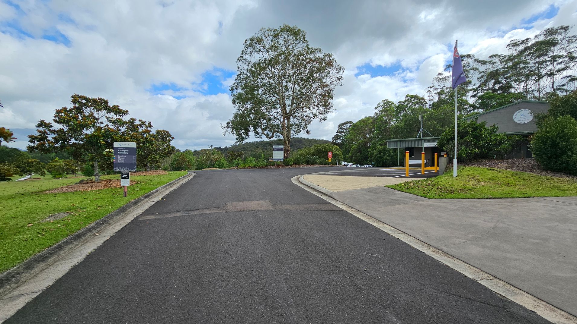 A paved road leads to a T-junction with a large tree in the centre. On the left, there's a grassy area with several smaller trees and a sign. On the right, a building with a green roof and a flagpole stands near a parking area. The background features a forested hill under a cloudy sky.