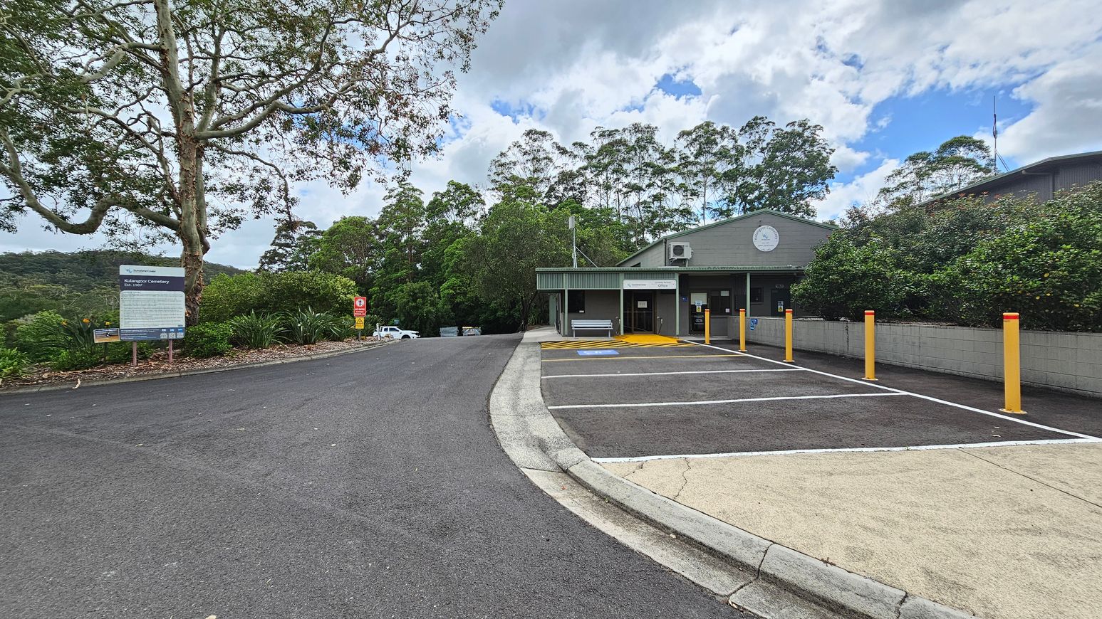 A paved road leads to a building on the right with a green roof and a clock on the facade. Yellow bollards line the edge of the parking area, which includes a disabled parking space. On the left, a large tree stands next to a sign with text, surrounded by greenery. In the background, dense trees cover the hillside. A white vehicle is partially visible further down the road.
