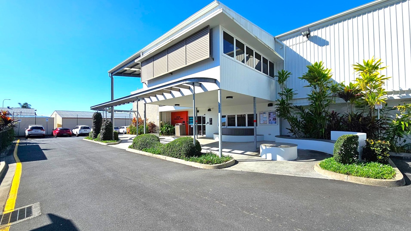 A modern building with a white exterior and large windows on the upper level. The entrance is sheltered by a metal awning supported by poles. To the right, there are landscaped areas with neatly trimmed bushes and tall plants. On the left, a driveway leads to a parking area with several cars. The ground is paved with asphalt, and the building's facade features a combination of horizontal and vertical lines.