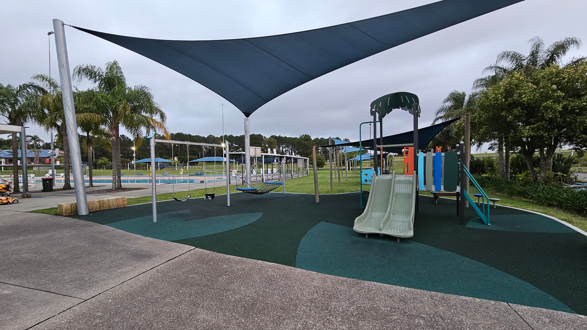 A playground with a slide and climbing structure on the right, featuring a green and blue colour scheme. In the centre, there are swings and a hammock under a large triangular shade sail. The ground is covered with a rubber safety surface in green and blue patterns. On the left, there is a concrete path and some palm trees. In the background, a fenced area with a pool and more shade structures is visible, along with more trees and grass.