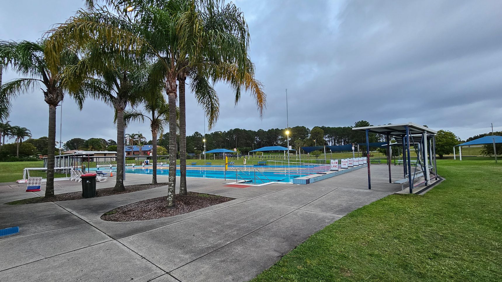 Concrete pathways surround a swimming pool in the centre, with lane markers visible. On the left, several tall palm trees stand beside a red bin. To the right, a metal-framed shelter with benches is situated on the grass. In the background, there are blue shade structures and a line of trees. The area is well-lit with outdoor lights.
