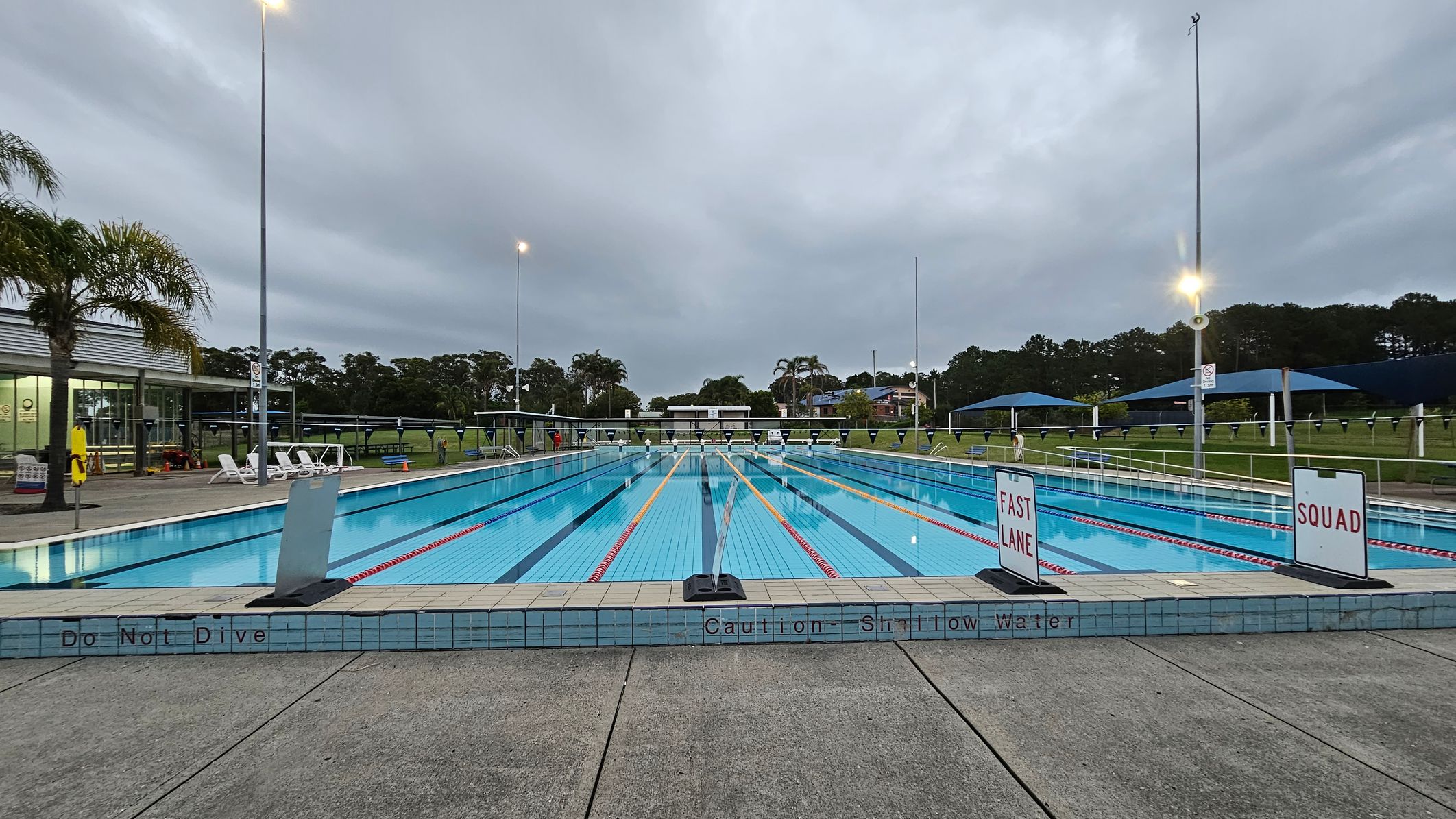 An outdoor swimming pool with multiple lanes, marked by red and white lane dividers. Signs reading "FAST LANE" and "SQUAD" are positioned at the pool's edge. On the left, there are white lounge chairs and a building with large windows. Palm trees and grassy areas surround the pool. Tall floodlights illuminate the area, and a grey, overcast sky is visible. The pool deck is made of concrete, with warnings like "Do Not Dive" and "Caution Shallow Water" written on the tiles.