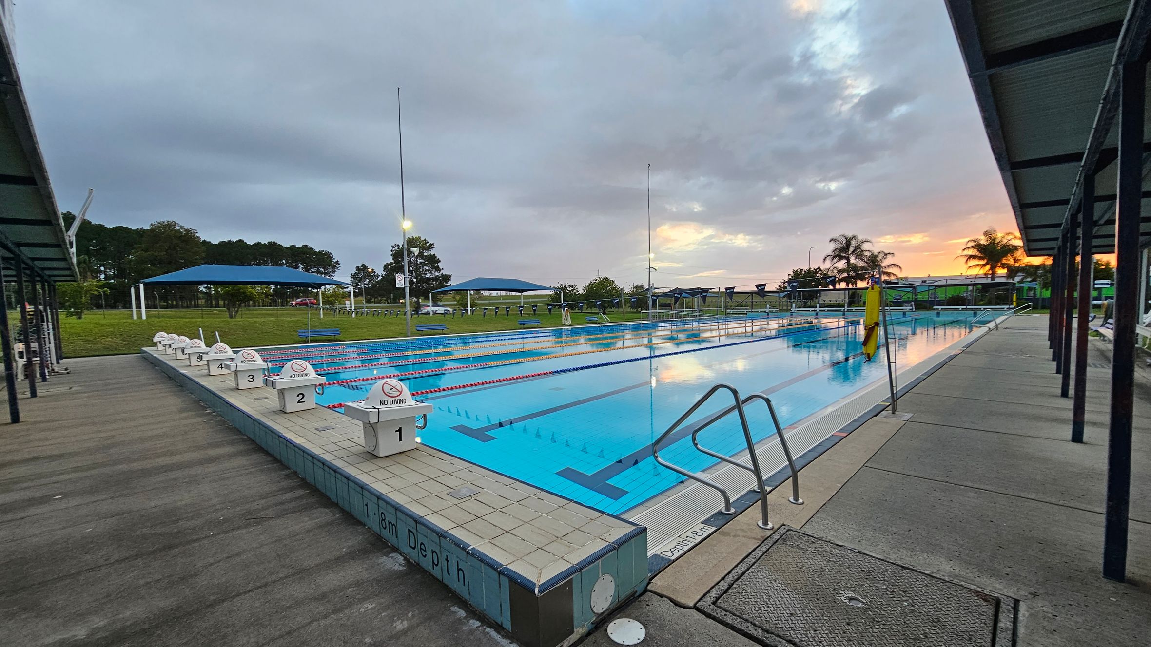 An outdoor swimming pool with multiple lanes, each marked with "No Diving" signs. The pool is surrounded by a concrete deck with metal handrails on the right. On both sides, there are covered walkways with green roofs. In the background, there are grassy areas with trees and blue shade structures. Palm trees are visible near the horizon, with a sunset casting a warm glow. The pool's depth is marked as 1.8 metres.