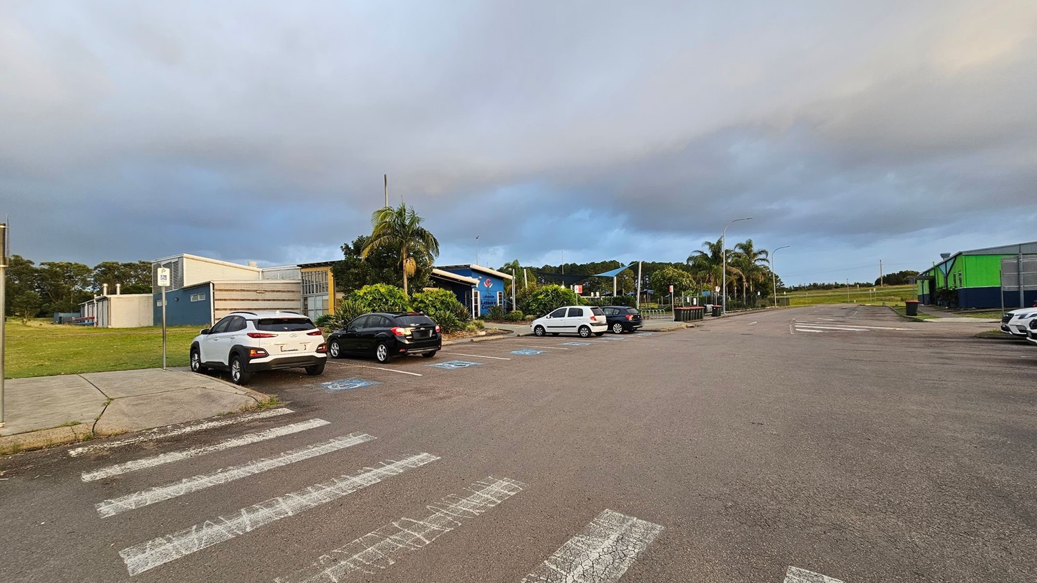 A paved road on the left with parked cars in the distance, lined with street lamps and trees, including palm trees. On the right, a grassy area is enclosed by a chain-link fence, featuring a structure with a blue roof and white poles. The background includes more trees and a few buildings.