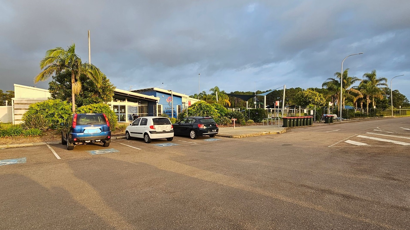 A car park with three cars parked in designated spaces on the left. A blue building with a sign reading "Leisure" is in the centre, surrounded by lush greenery and palm trees. On the right, there are several rubbish bins and a few more palm trees lining the road. The ground is paved, and there are streetlights along the road.
