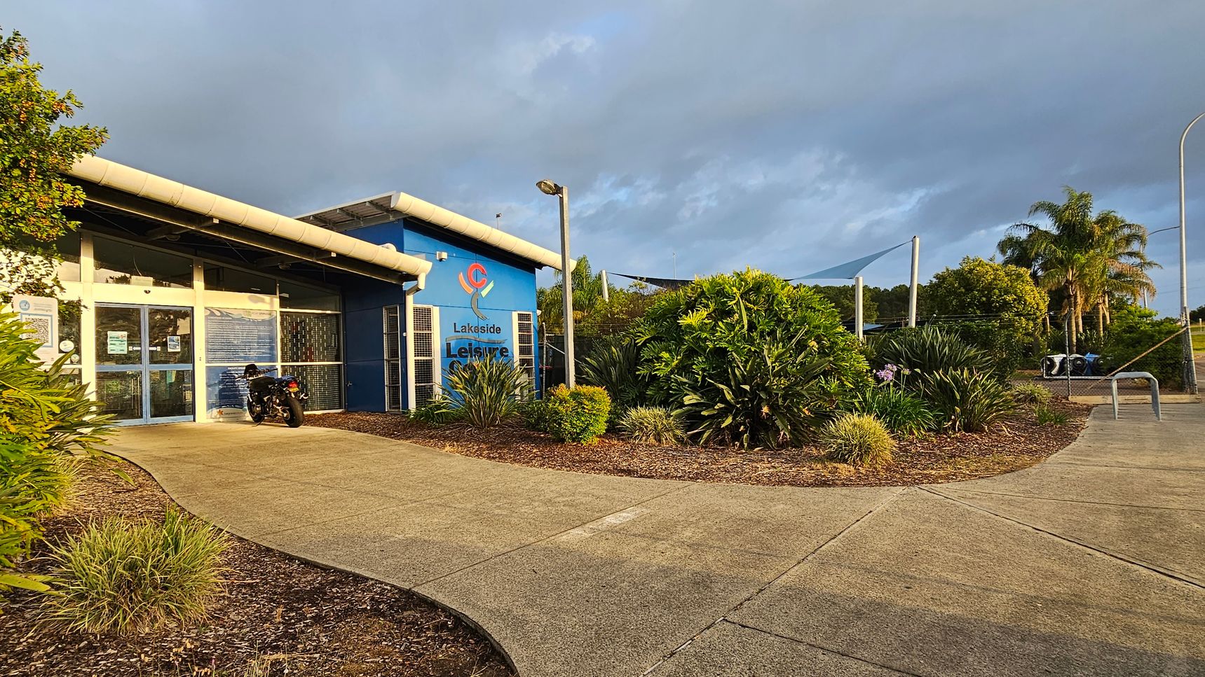 A leisure centre with a blue exterior is on the left, featuring a glass entrance and a motorcycle parked nearby. The building is surrounded by landscaped greenery, including bushes and small trees. A concrete path leads to the entrance, curving through the garden. On the right, there are more plants and a few palm trees in the background. A metal bike rack is visible near the path. The area is well-lit by a streetlamp.
