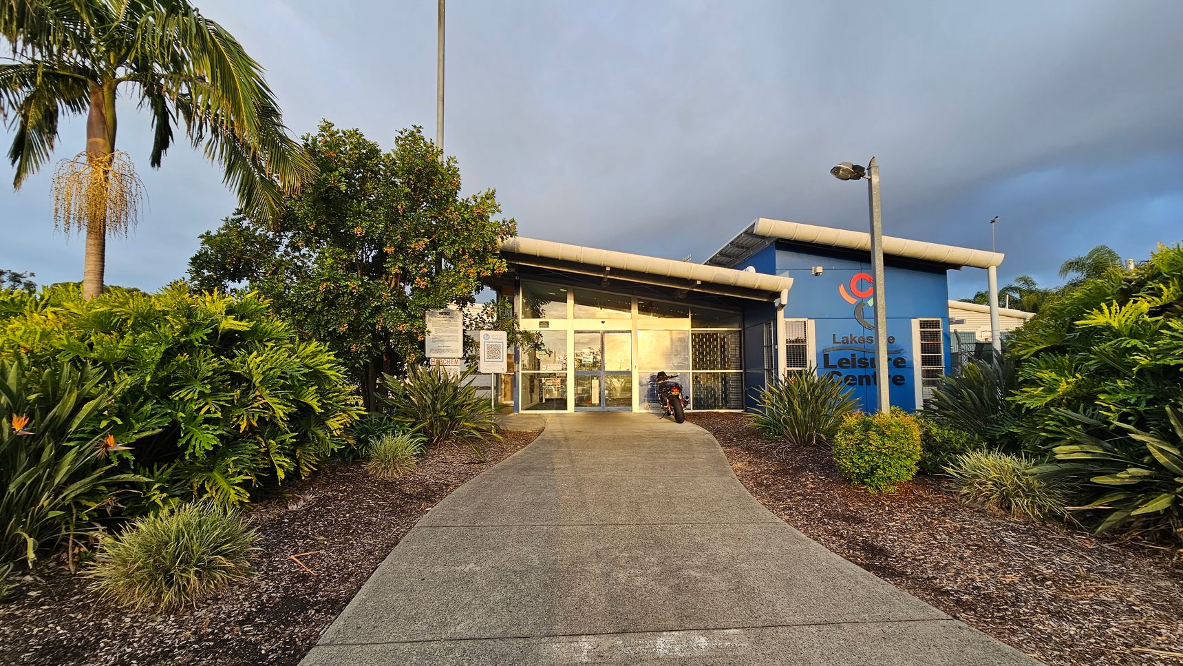 A building with a glass entrance is in the centre, with a path leading up to it. On the right, there's a blue wall with the words "Lakeside Leisure Centre" and a logo. A motorcycle is parked near the entrance. The foreground has a variety of green plants and shrubs, with a palm tree on the left. Two tall lamp posts are positioned near the entrance. The building has a slanted roof, and the area is surrounded by lush greenery.