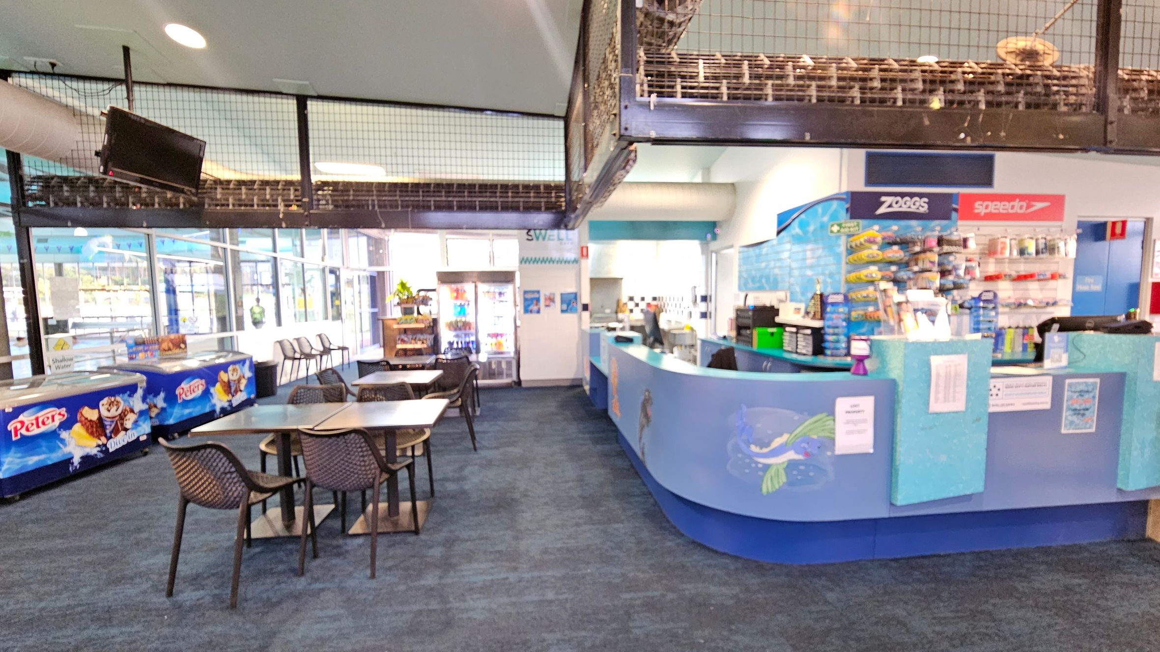A café area with a blue-themed counter on the right, displaying various swimming accessories and snacks. Behind the counter are shelves with products. In the centre, there are tables and chairs with a woven design. On the left, there are ice cream freezers branded "Peters" and a wall of windows. Above, a television is mounted near a mesh barrier. The floor is carpeted in a dark blue pattern.