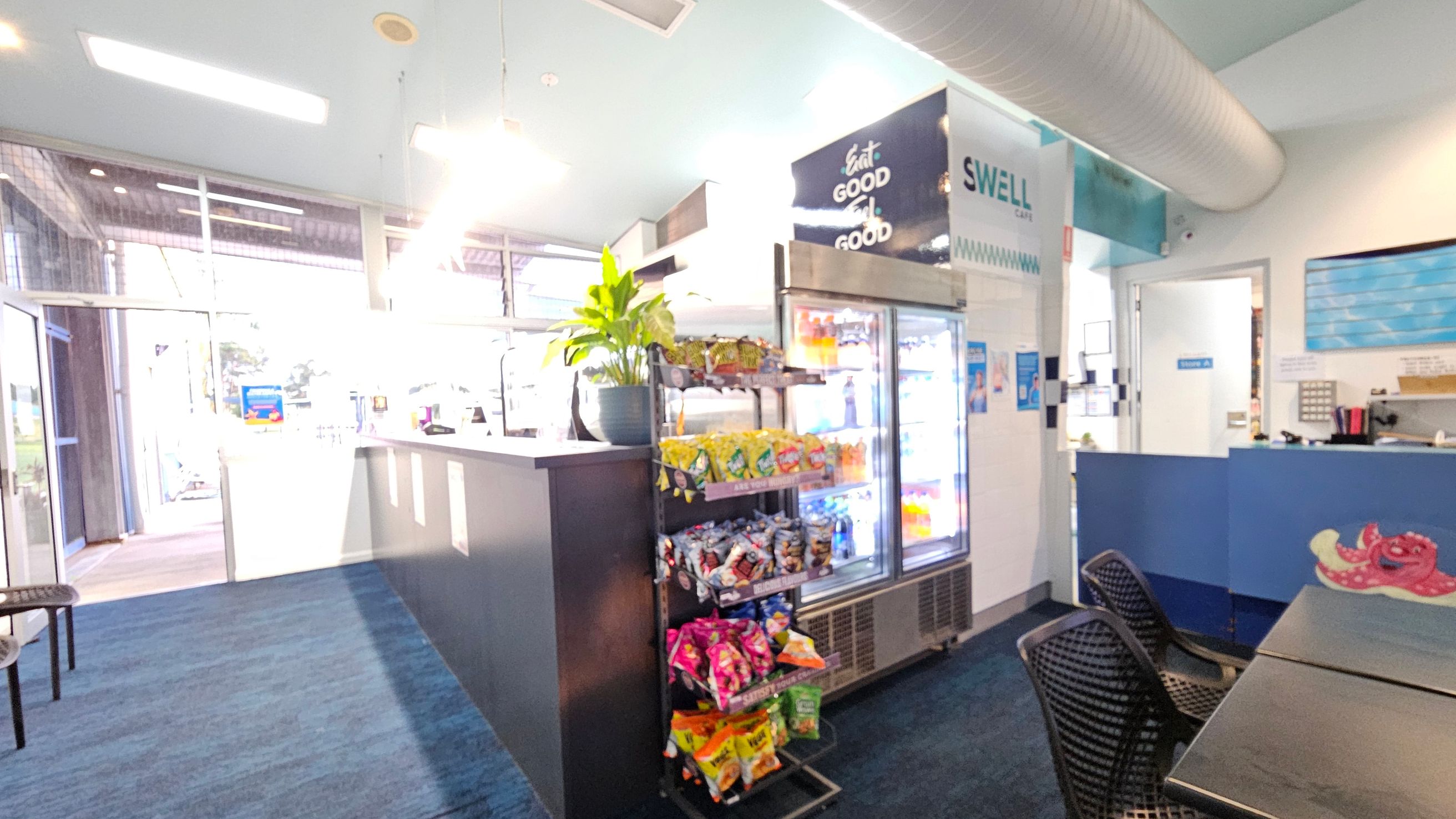 Inside a café with a blue carpeted floor. On the left, there's a glass entrance with natural light streaming in. In the centre, a counter with a plant on top and a snack display below. Behind it, a refrigerator stocked with drinks. On the right, a seating area with black chairs and tables. Above, a large duct runs along the ceiling. A sign reads "SWELL Café" and a colourful octopus design is visible on the counter.