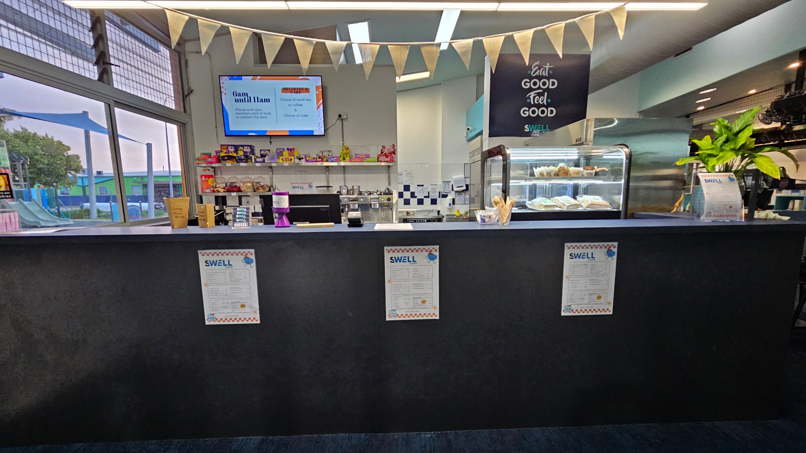 A café counter with a dark surface and three menus displayed on the front. Behind the counter, there is a display case with pastries and sandwiches. A plant is on the right side. Above, a string of triangular flags hangs, and a sign reads "Eat Good Feel Good." A TV screen on the left shows menu options. Large windows on the left reveal an outdoor area with playground equipment. The ceiling has modern lighting fixtures.