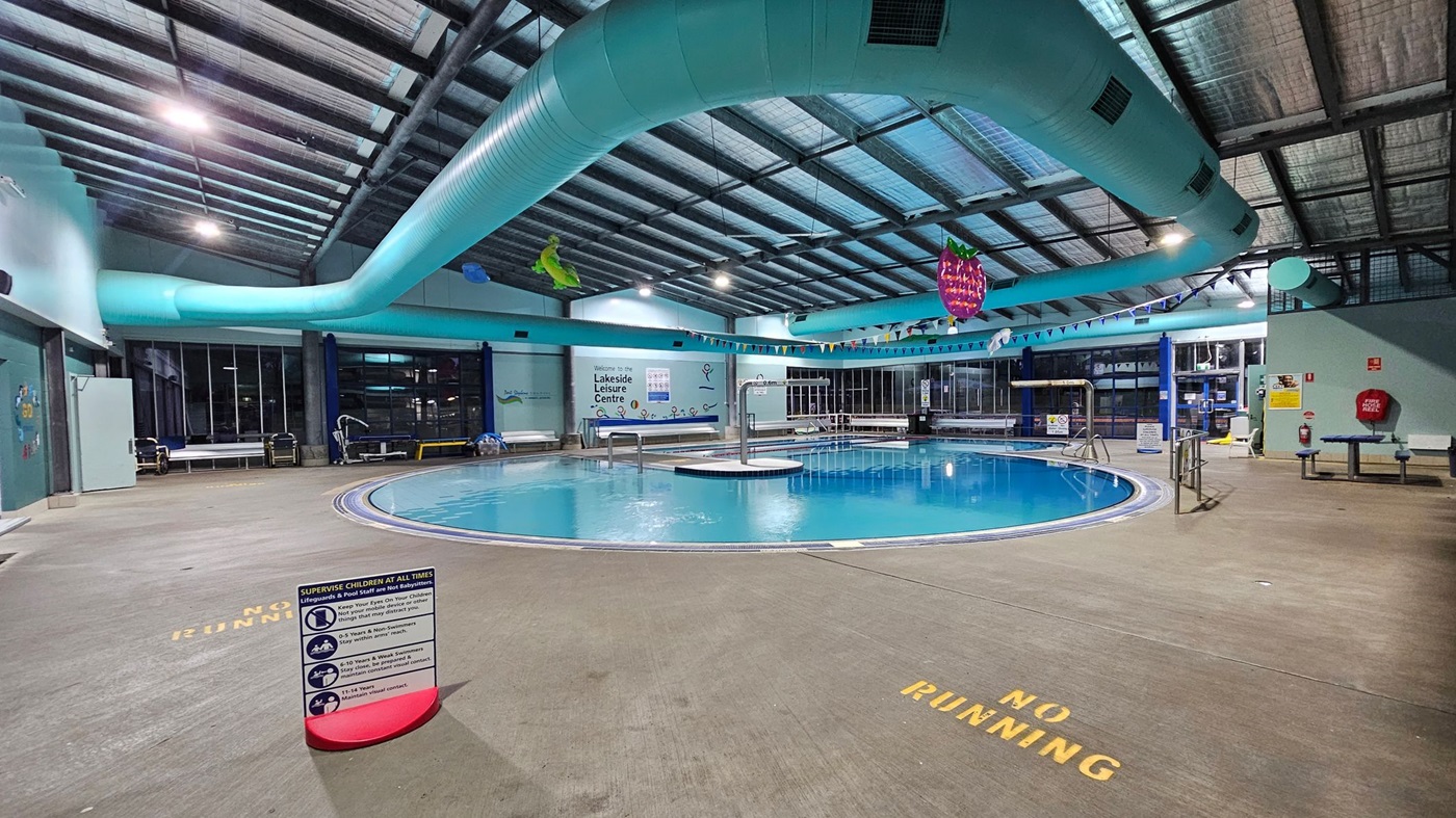 A glass entrance to a swimming pool area with blue-framed doors. On the left, a board displays photos and names of swim teachers at the Lakeside Leisure Centre. Below it, a white wire basket with a red sale sign. On the right, a vertical banner promoting "Float to Survive with GOswim" stands next to the door. Through the glass, a pool with turquoise pipes and aquatic-themed decorations is visible. The floor is grey, and the ceiling has exposed beams and lights.