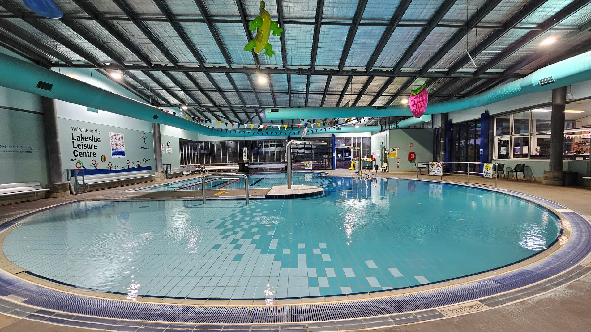 Indoor swimming pool with a curved edge in the foreground, surrounded by a tiled floor. The ceiling is high with exposed beams and a large inflatable turtle and strawberry hanging from it. On the left, a wall displays "Welcome to the Lakeside Leisure Centre" with benches below. The right side has large windows and a seating area with chairs. A blue duct runs along the ceiling, and colourful bunting is strung across the pool area.