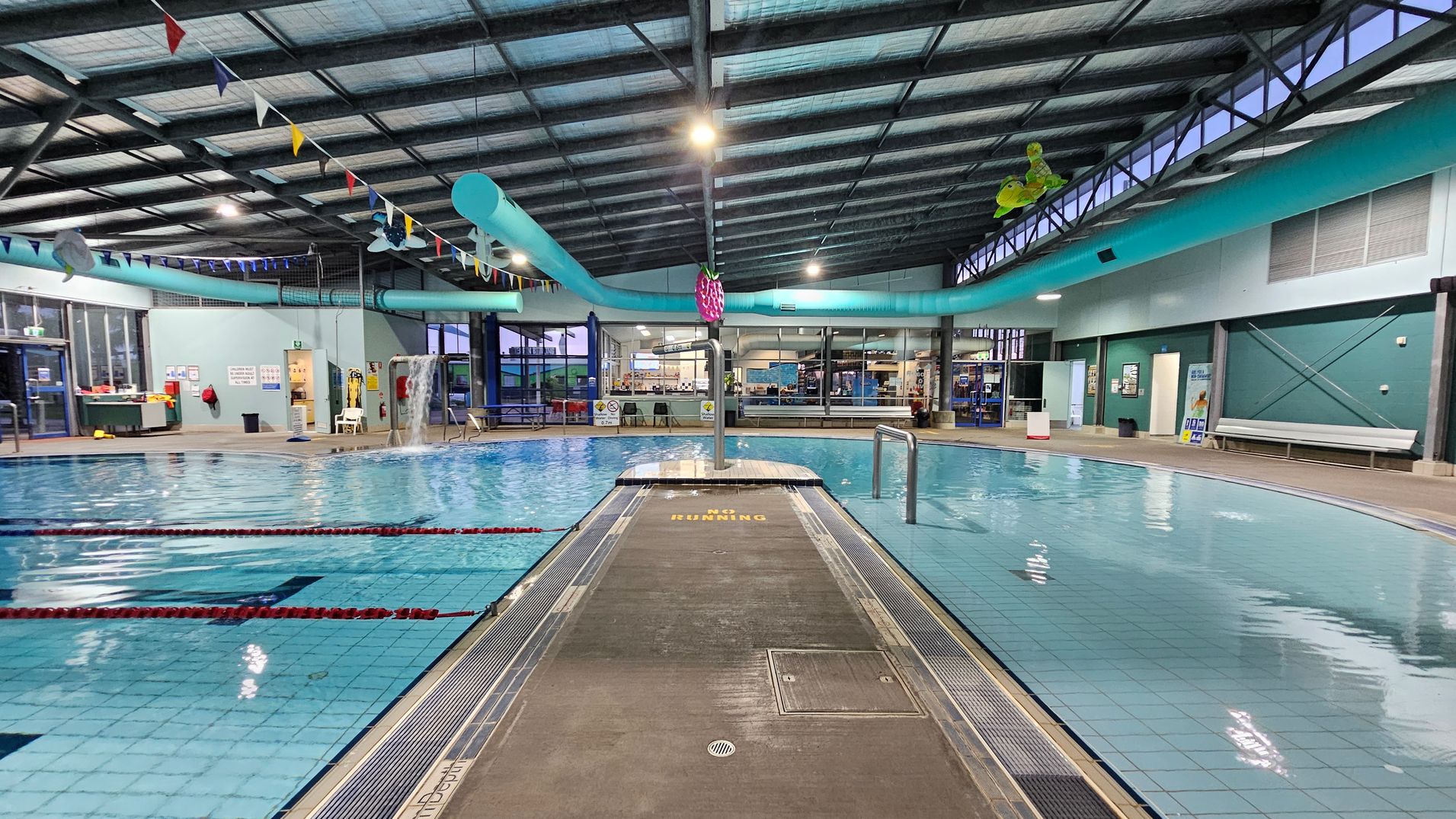 Indoor swimming pool with a tiled floor and a central walkway marked "No Running." On the left, there are lanes with red lane dividers. The right side features a shallow area with a small waterfall. The ceiling has exposed beams and colourful bunting. In the background, there are large windows and a seating area with benches. A blue water slide curves above the pool, and there are various safety and informational signs on the walls.