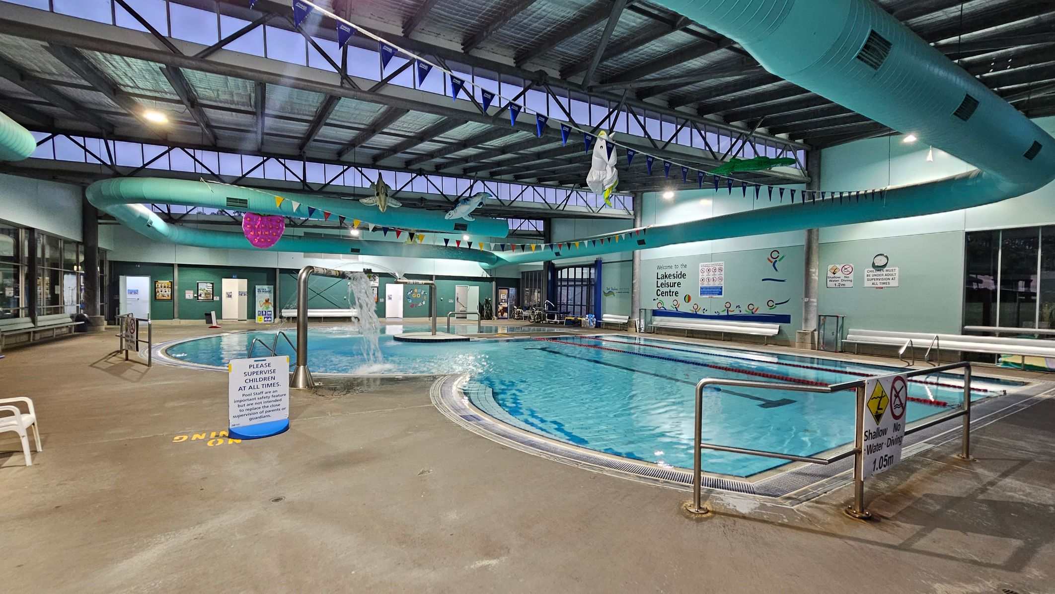 Indoor swimming pool area with a large pool in the centre. The pool has a water feature and is surrounded by a concrete deck. On the left, there are benches and a sign instructing supervision of children. The right side has a railing with a sign indicating shallow water. The ceiling features exposed beams and a large turquoise ventilation duct. Colourful bunting and inflatable decorations hang above. Walls have informational signs and windows.