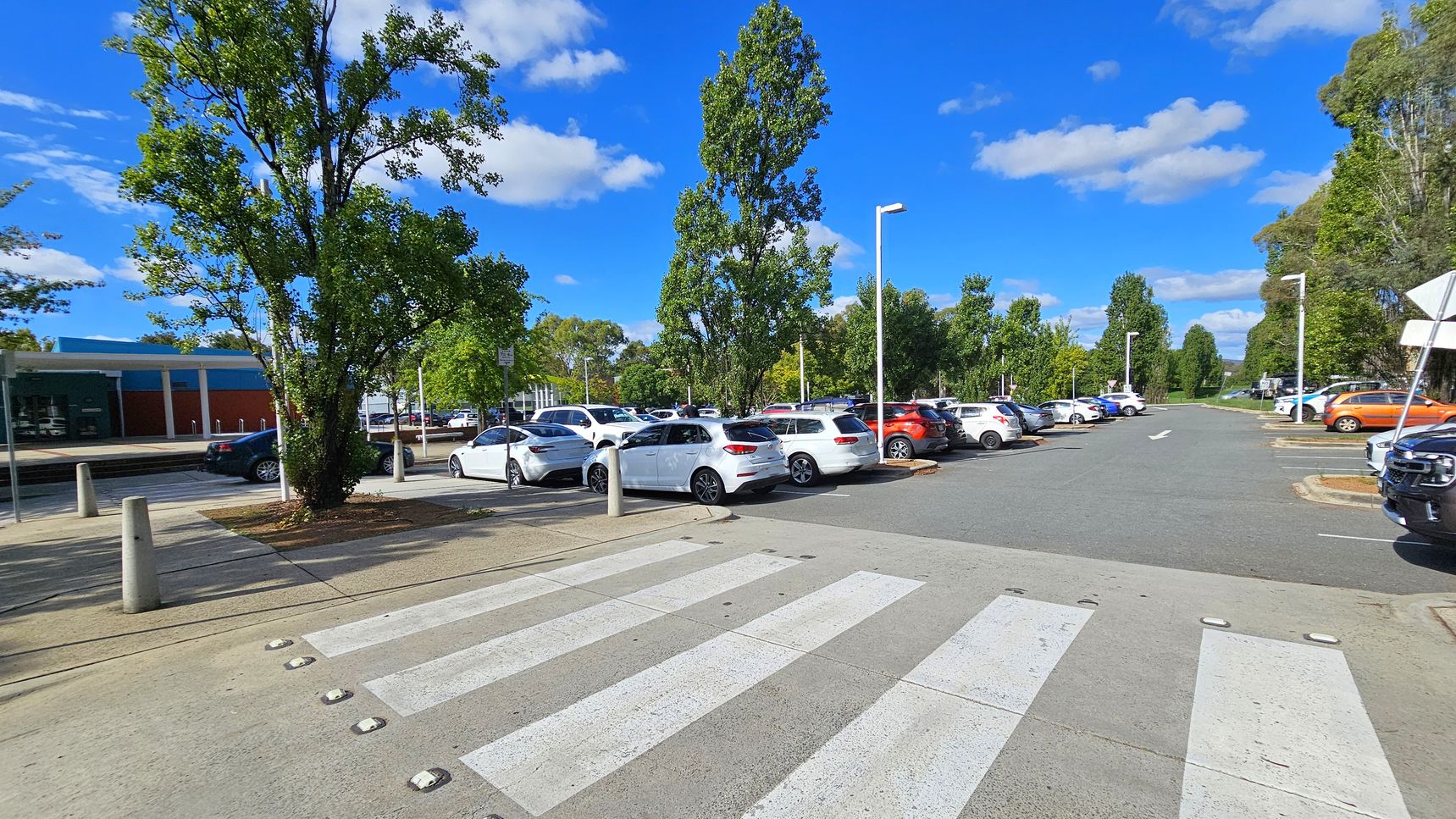 A car park with a zebra crossing in the foreground. On the left, there is a large tree and a building with a blue and red facade. Several cars are parked, mostly white, with a few in other colours like orange. Tall trees line the parking area, and street lamps are visible. The road extends into the distance, flanked by more trees and greenery. The sky is clear with a few clouds.