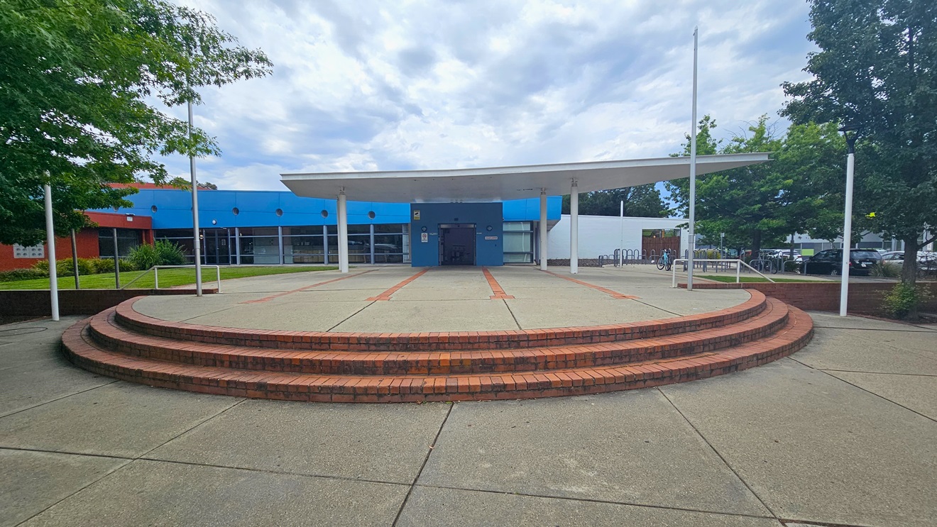 A circular, tiered brick platform is in the foreground, leading up to a covered entrance. The building has a blue facade with large windows and a flat roof supported by white pillars. On the left, there are green trees and a red section of the building. On the right, more trees and a glimpse of parked cars are visible. The ground is paved with concrete, and the sky is overcast.