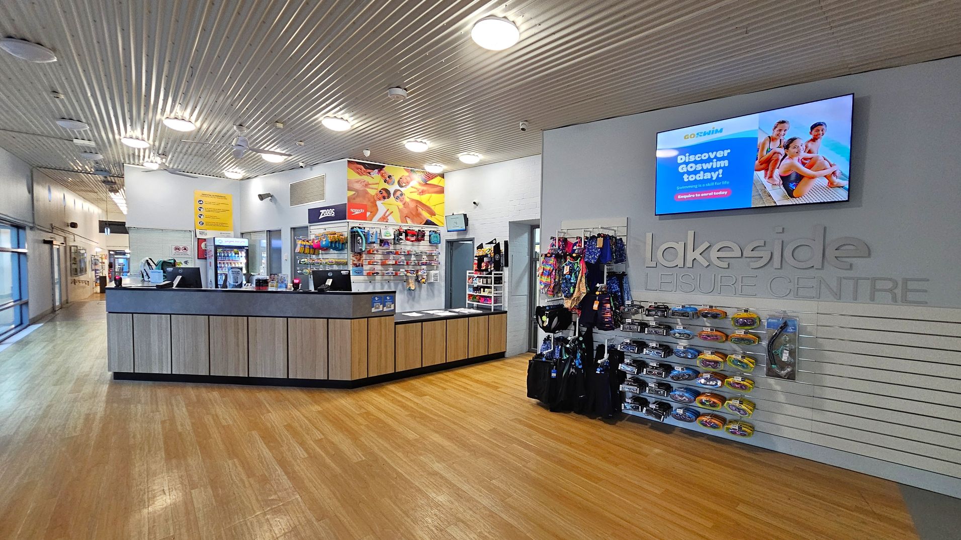 Reception area of a leisure centre with a wooden floor and a corrugated metal ceiling. On the left, a reception desk with computers and a display of swimming accessories. Behind the desk, a colourful poster and a vending machine. On the right, a wall-mounted display screen above the words "Lakeside Leisure Centre" and racks of swim gear. The background features a corridor with glass windows.