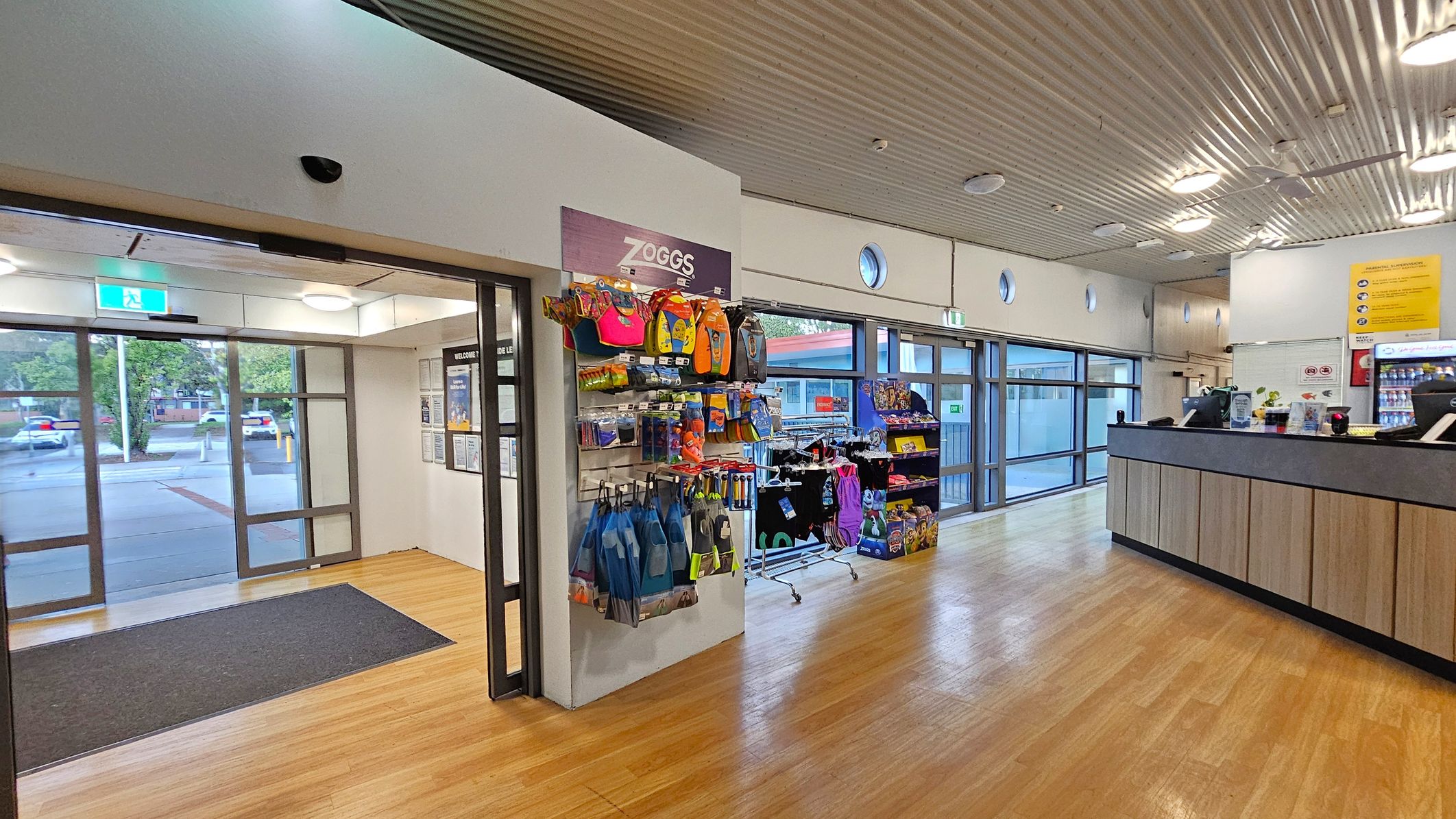 A spacious interior with a wooden floor and a corrugated metal ceiling. On the left, there is a glass entrance with a black mat and a view of parked cars outside. In the centre, a display rack features colourful swim gear and accessories. To the right, a wooden counter with a black top has a cash register and promotional materials. Large windows line the wall, allowing natural light to fill the space.