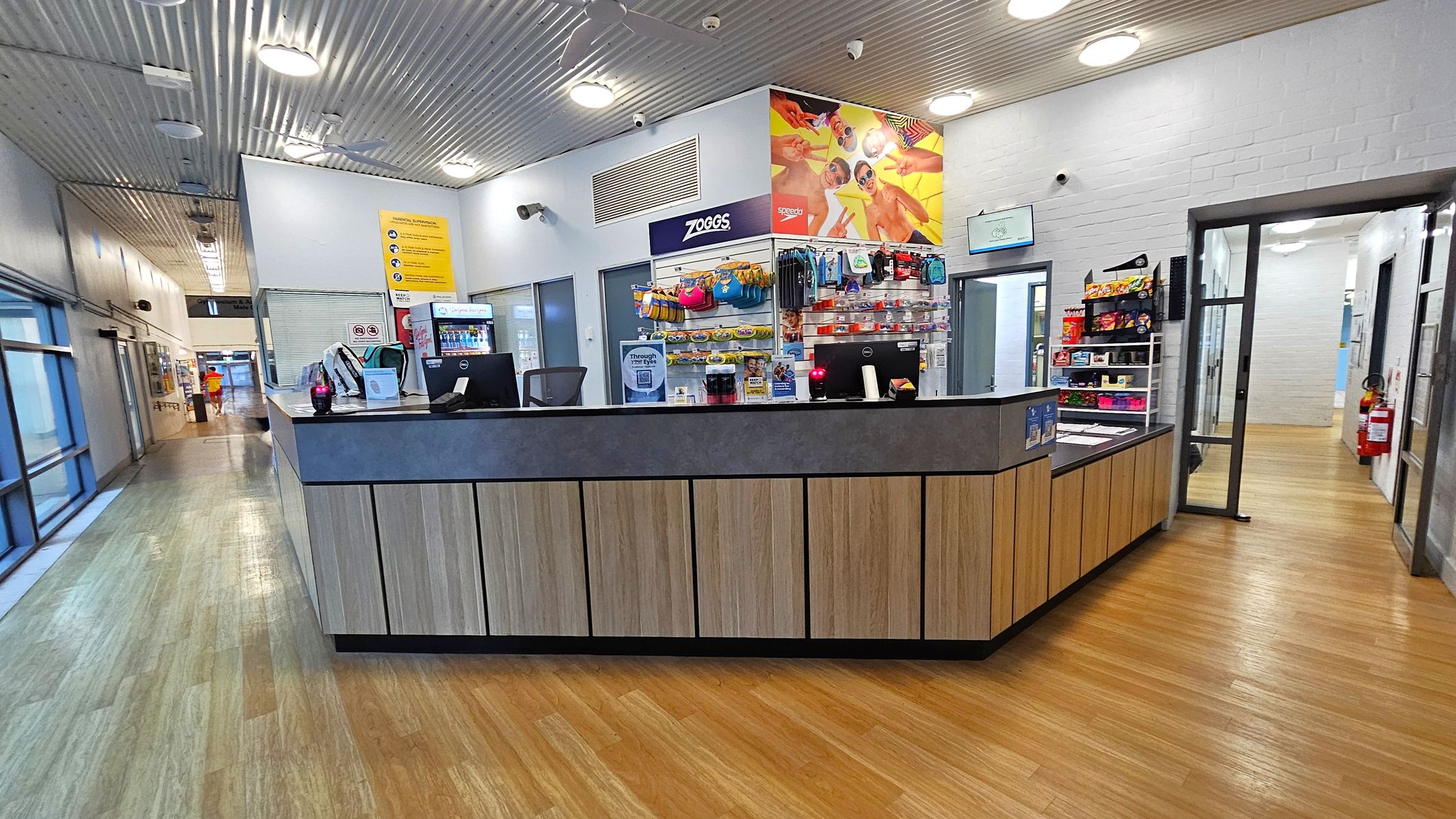 A reception area with a wooden counter in the centre, featuring two computer monitors and various items like a backpack and a drink bottle. Behind the counter, shelves display swimming gear and snacks. The floor is wooden, and the ceiling has a metallic finish with circular lights. To the left, a corridor with glass doors and windows leads to another area. On the right, a doorway opens to a hallway with a fire extinguisher mounted on the wall.