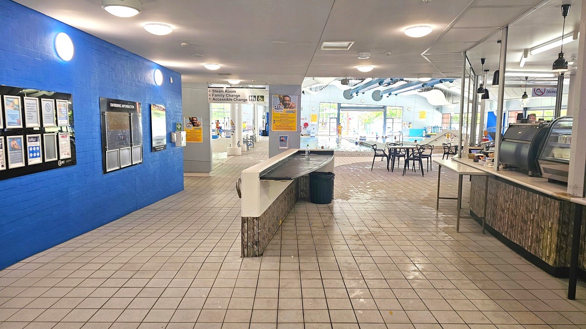 A spacious indoor area with a tiled floor and ceiling lights. On the left, a bright blue wall displays various notices and information boards. In the centre, a long counter with a bin underneath leads towards a pool area visible in the background. On the right, a café counter with a glass display case and hanging lights is adjacent to a seating area with black chairs and tables. The pool area in the background features large windows and visible ductwork on the ceiling.