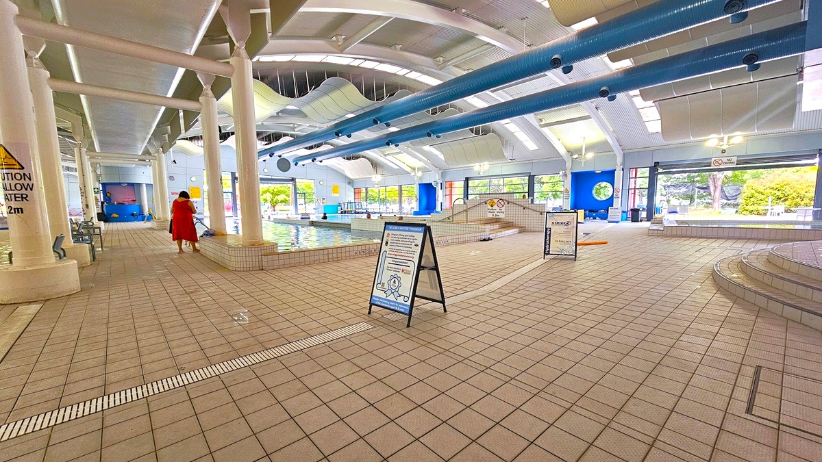 Indoor swimming pool area with tiled flooring and a high ceiling featuring blue pipes. On the left, a person in a red outfit walks towards the pool, which is surrounded by white columns. In the centre, a signboard stands on the floor. The right side has steps leading to another section of the pool. Large windows in the background allow natural light to illuminate the space, and there are caution signs around the pool area.