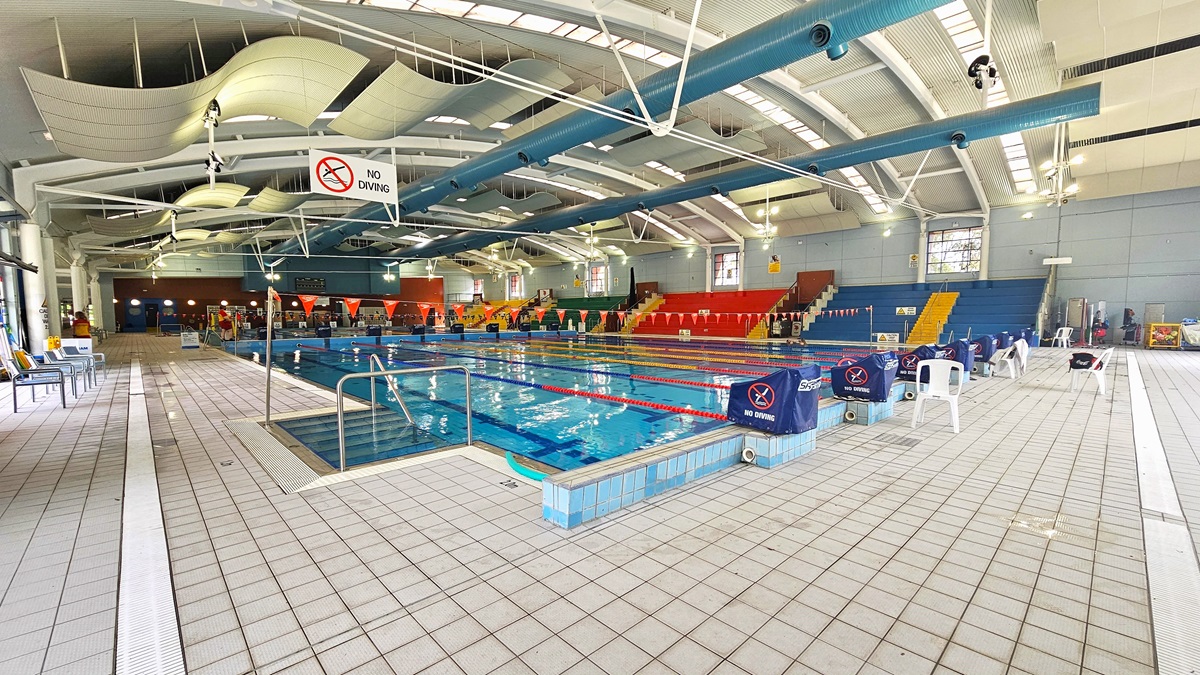 An indoor swimming pool with multiple lanes, surrounded by a tiled deck. On the left, there are chairs and a "No Diving" sign hanging from the ceiling. The right side features more chairs and a few white plastic chairs. In the background, there are colourful spectator stands in red, yellow, and blue. The ceiling has large, curved white panels and exposed beams. The pool area is well-lit with natural light coming through the windows.