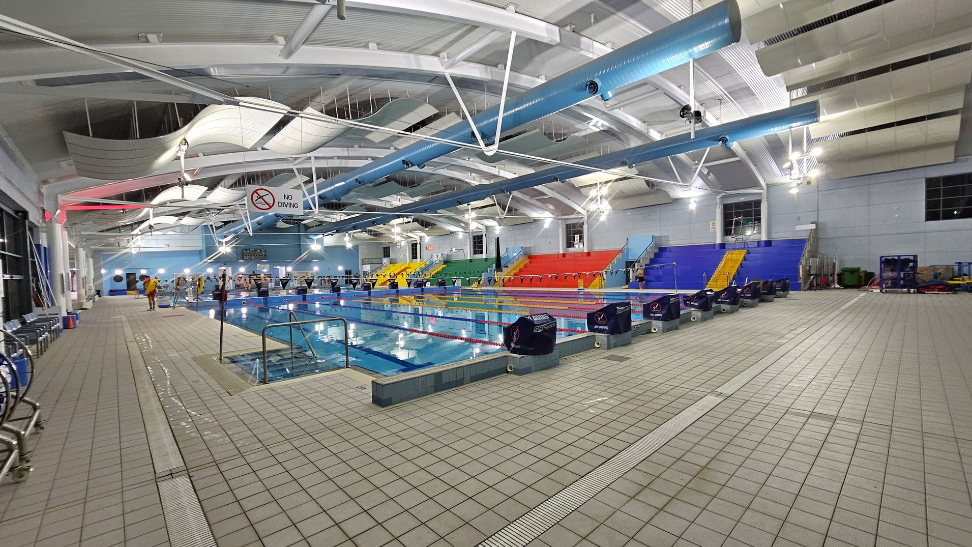 An indoor swimming pool with multiple lanes, surrounded by a tiled floor. On the left, there are chairs and a "No Diving" sign. In the middle, the pool is lined with lane dividers and starting blocks. On the right, there are colourful spectator seats in blue, red, yellow, and green. The ceiling features exposed beams and ductwork, with overhead lighting illuminating the area.