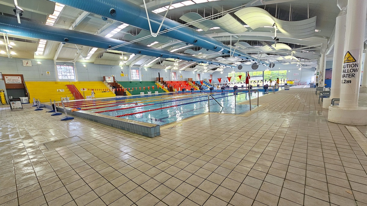 Indoor swimming pool with lanes marked by red and white ropes. On the left, there are yellow and green spectator seats with red stairs. The right side has a tiled pool deck with a caution sign on a pillar. The ceiling features large blue pipes and white curved panels. The floor is covered with light-coloured tiles. Bright overhead lighting and windows provide illumination.