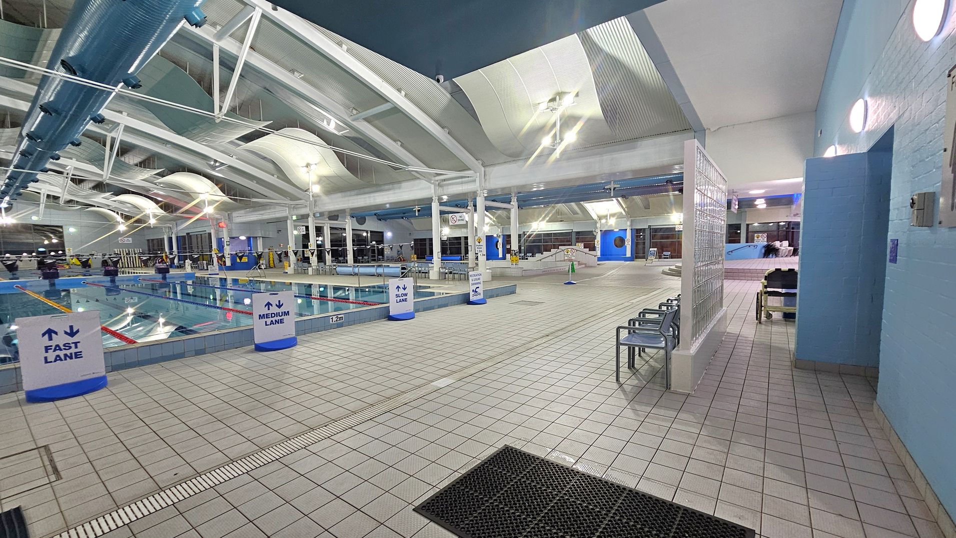 Indoor swimming pool area with lanes marked as fast, medium, and slow. The pool is on the left, with clear water and lane dividers. The floor is tiled in a light colour, with a black mat near the entrance on the right. Several metal chairs are positioned along the wall on the right. The ceiling is high with white beams and large, curved light fixtures. The walls are painted light blue, and there are glass blocks near the entrance. Bright overhead lighting illuminates the space.