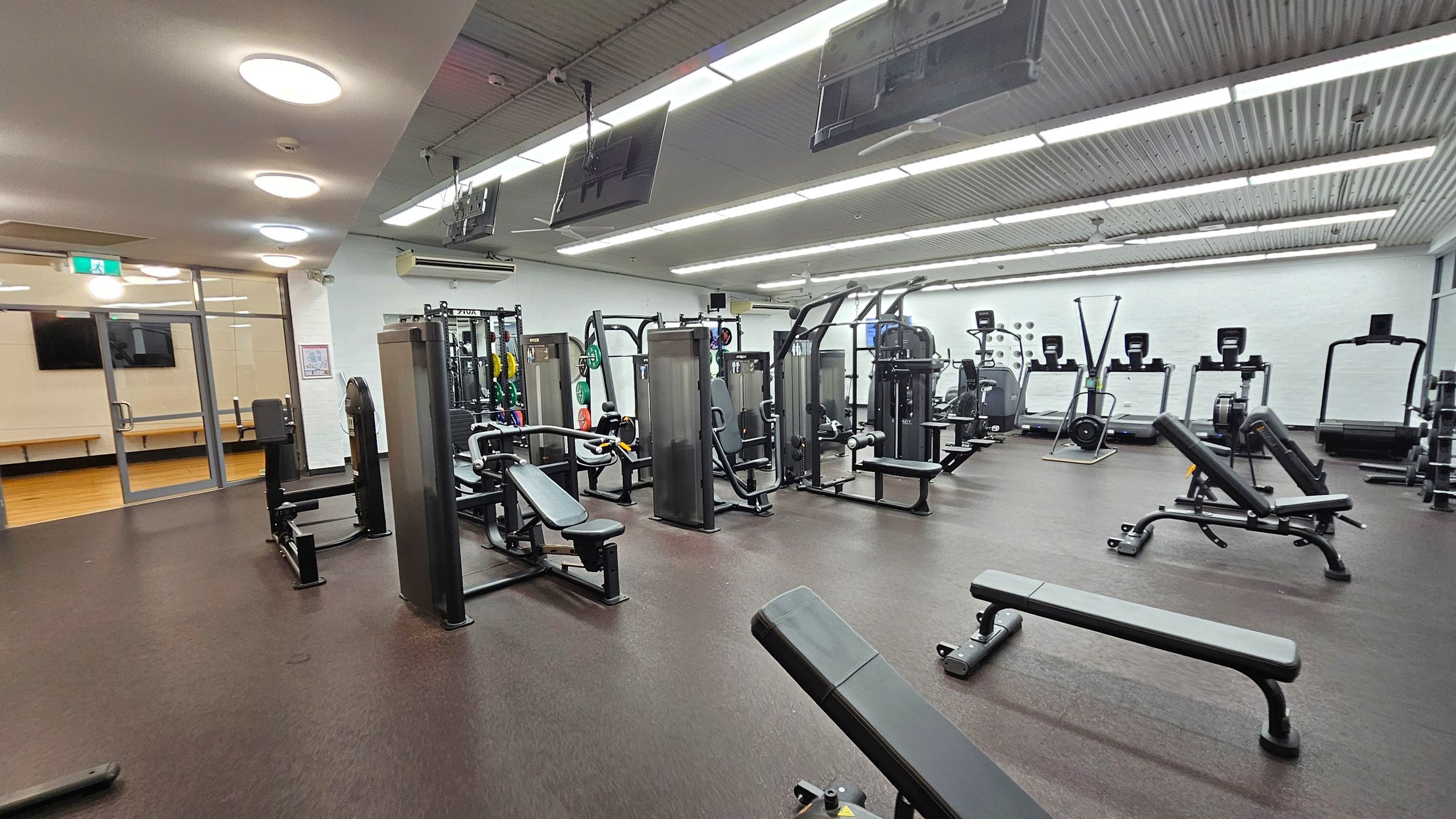 A spacious gym with various exercise equipment. On the left, there are weight machines and benches. In the centre, more weight machines and benches are arranged neatly. On the right, treadmills and rowing machines are lined up against the wall. The floor is dark and rubberised, while the ceiling features bright, recessed lighting. Large windows and a glass door are visible on the far left, leading to another room with wooden flooring.