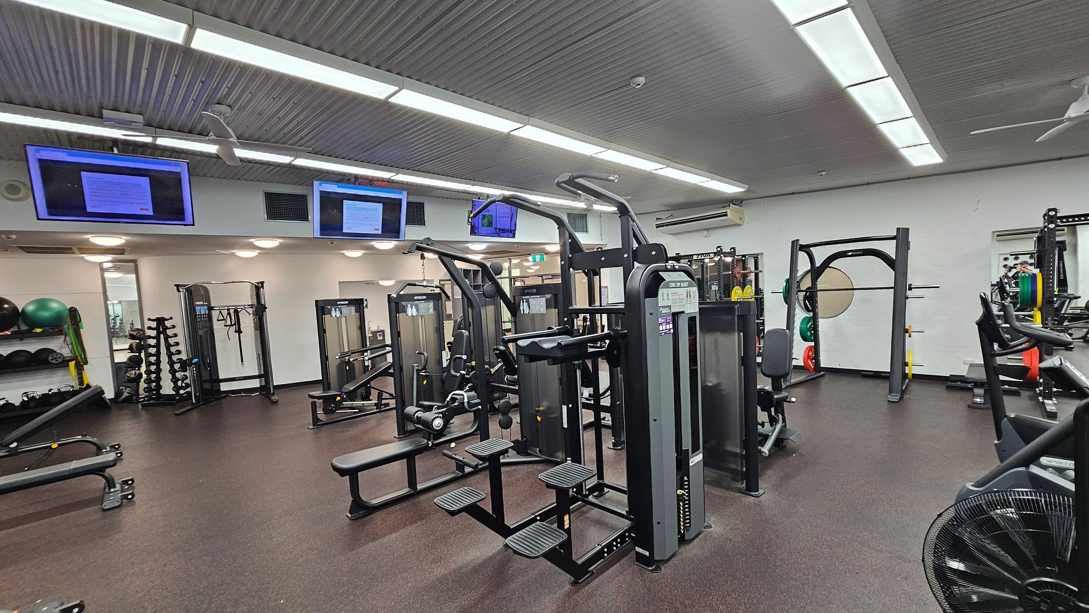 A gym with various exercise equipment. In the centre, there are weight machines and benches. On the left, shelves hold exercise balls and resistance bands, with a rack of dumbbells nearby. On the right, a squat rack with weight plates is visible. The ceiling has fluorescent lights and mounted screens displaying information. The floor is covered with dark rubber matting.