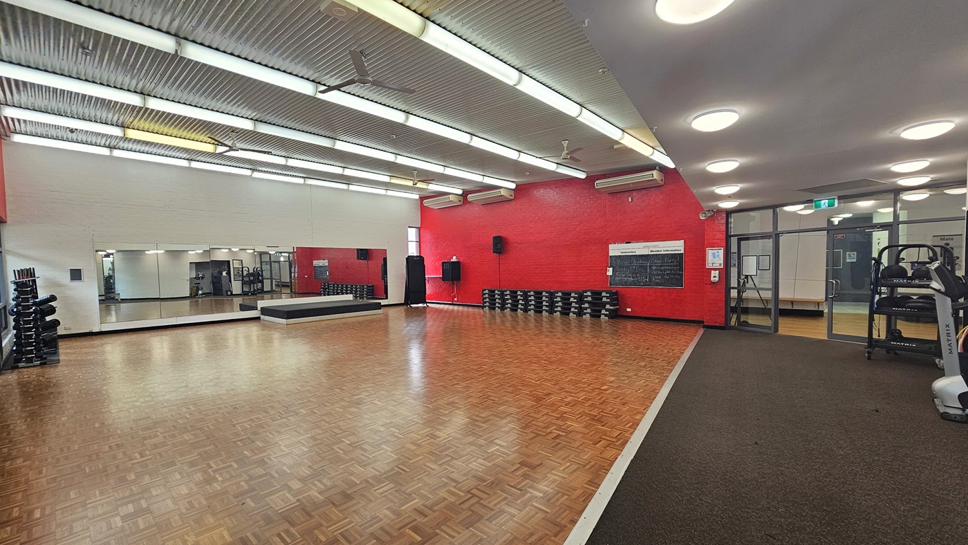 A spacious gym room with a wooden parquet floor. On the left, there are stacks of dumbbells and a large mirror covering the wall. The back wall is painted red and features a row of stacked step platforms and a blackboard. On the right, there is a treadmill and a glass door leading to another room. The ceiling is metallic with fluorescent lights, and the floor transitions to a carpeted area near the treadmill.