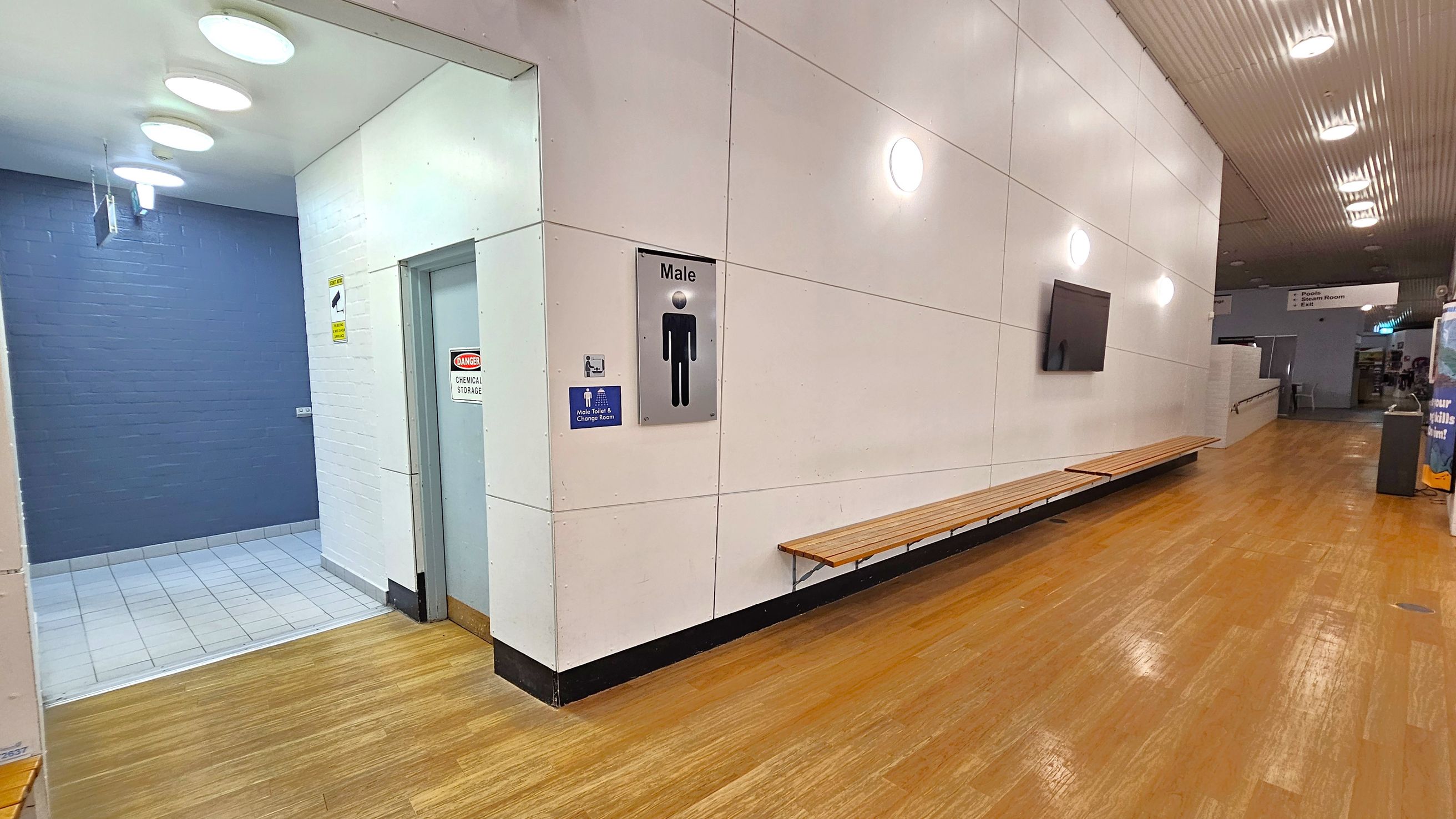 A corridor with wooden flooring and a white panelled wall on the right. A long wooden bench is attached to the wall. A sign indicating a male change room is visible. On the left, a tiled area leads to a door marked "Danger Chemical Storage." The ceiling has circular lights, and further down the corridor, there are signs and a vending machine.