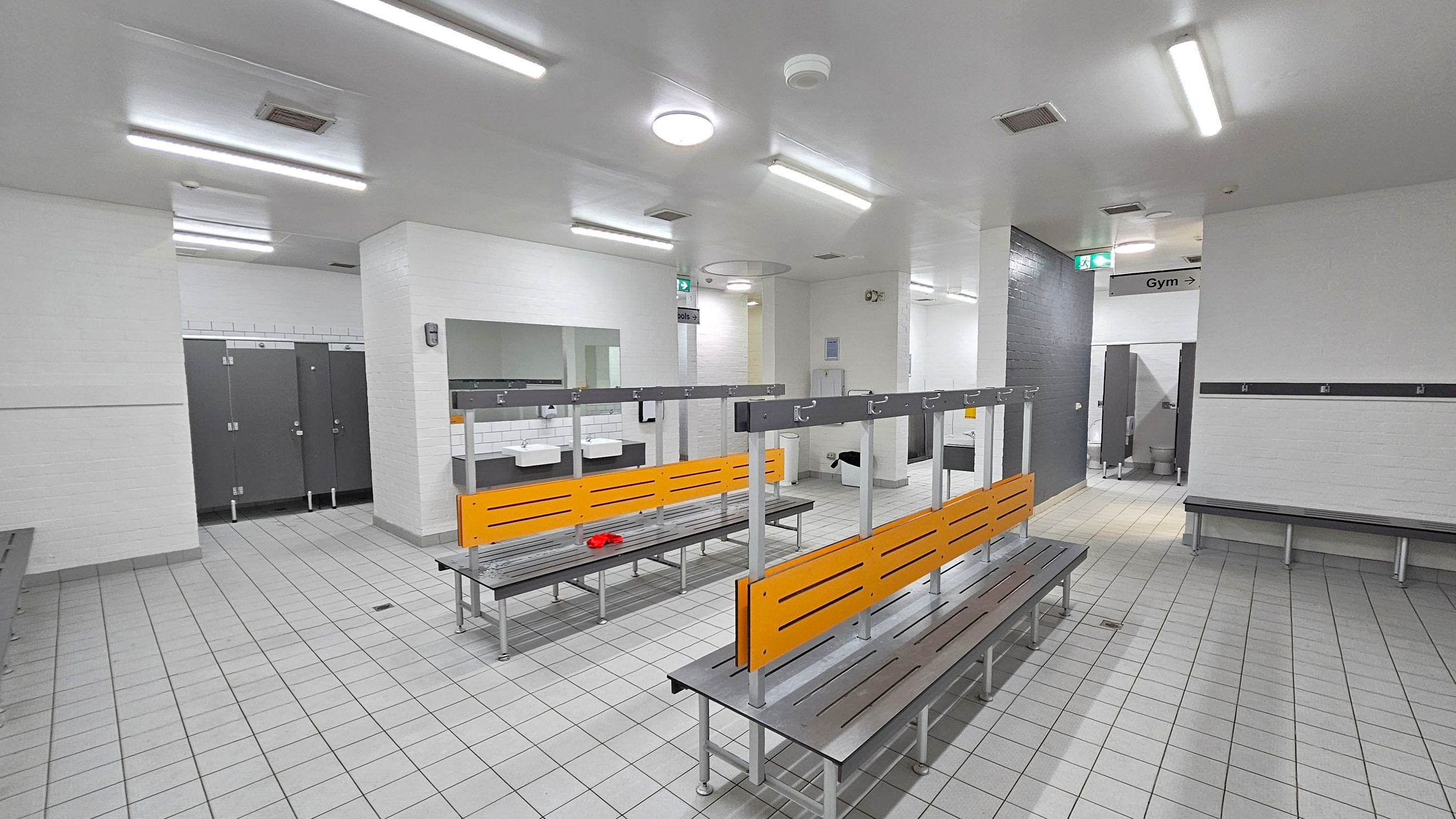 A spacious locker room with grey tiled flooring and white walls. In the centre, there are benches with orange backrests and metal hooks above. To the left, there are several closed grey lockers. In the background, sinks with mirrors are visible. On the right, a sign indicates the direction to the gym. The ceiling is white with fluorescent lights. A red item is placed on one of the benches.