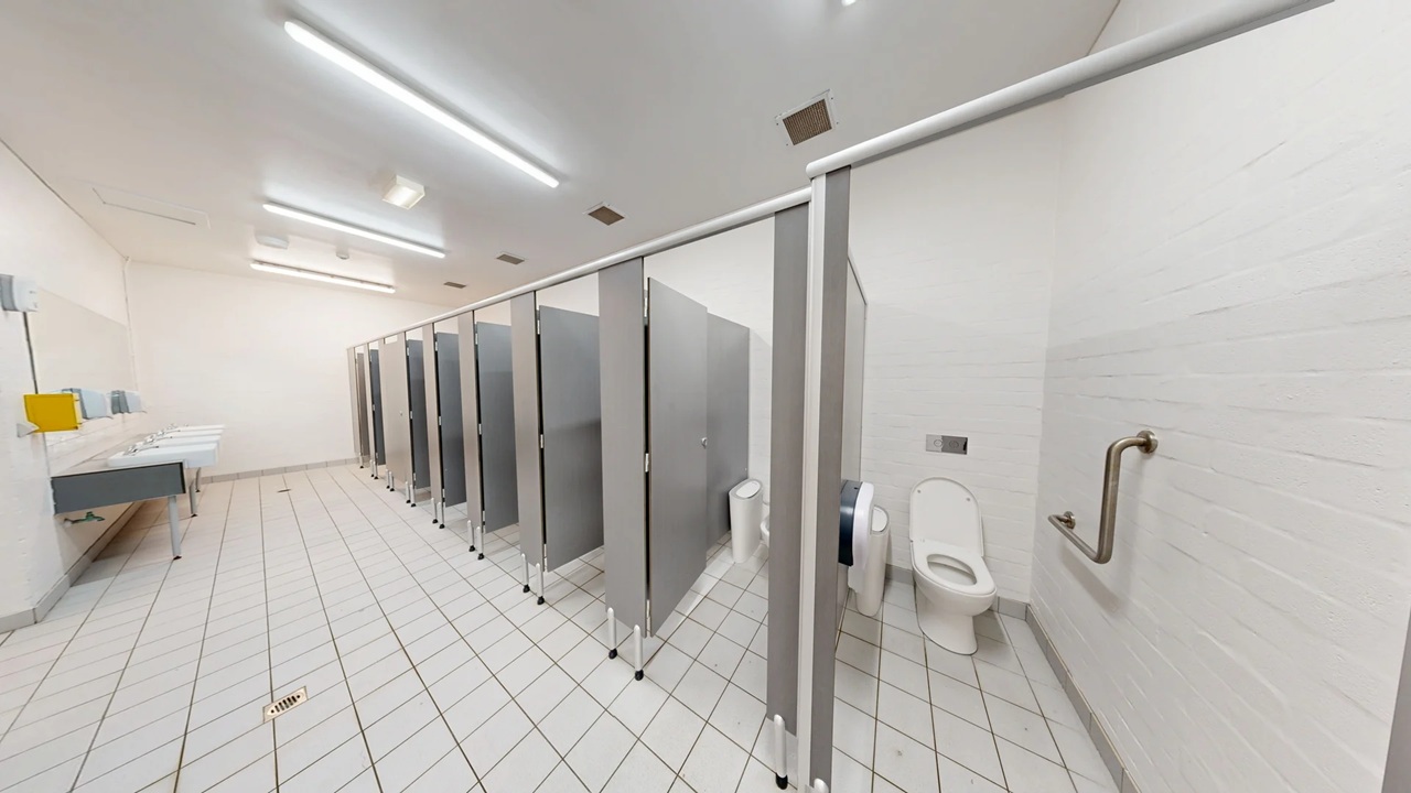 A public restroom with a row of toilet cubicles on the right, each with grey doors and partitions. The first cubicle door is open, revealing a white toilet and a grab bar on the wall. On the left, there are sinks with a yellow soap dispenser and a hand dryer. The floor is covered with white tiles arranged diagonally, and the ceiling has fluorescent lights. The walls are plain white, contributing to a clean and minimalistic appearance.