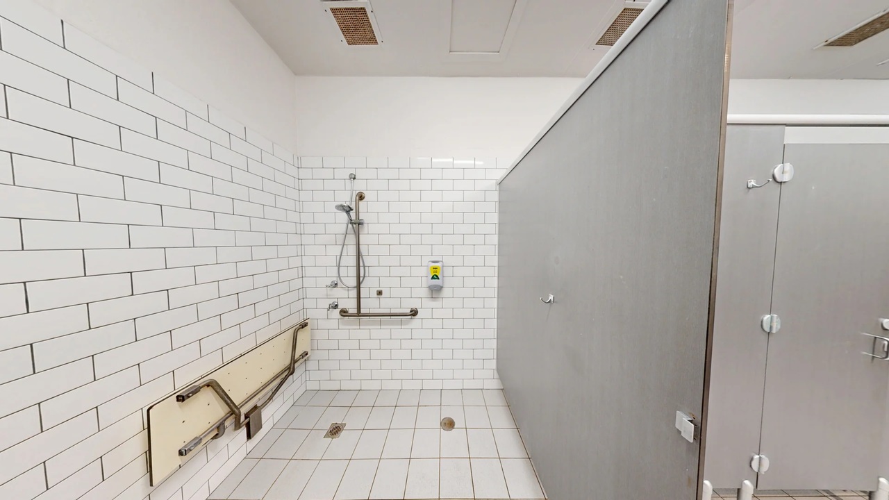 A bathroom with white subway tiles on the walls and floor. On the left, there is a foldable bench with metal support bars. In the centre, a showerhead is mounted on the wall with a grab bar below it. A soap dispenser is also visible. On the right, a grey partition separates the shower area from another section. The ceiling is plain white with ventilation panels.